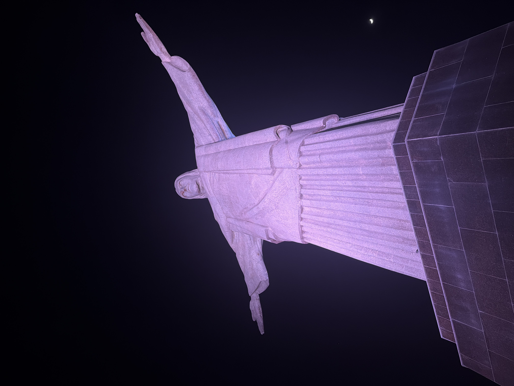 The image shows the Christ the Redeemer statue in Rio de Janeiro, Brazil, at night. The iconic Art Deco statue of Jesus Christ is illuminated with purple light, casting a dramatic glow against the dark sky. The statue stands on a pedestal made of dark, stone blocks, arranged in a tiered structure. The angle of the photograph is from below, looking up at the statue, emphasizing its imposing scale. In the upper right corner of the image, a sliver of a crescent moon is visible in the night sky. The statue's arms are outstretched, and its robe drapes down its body. The texture of the stone is visible, highlighting the details of the sculpture. The dark, cloudless sky provides a stark contrast to the illuminated monument.