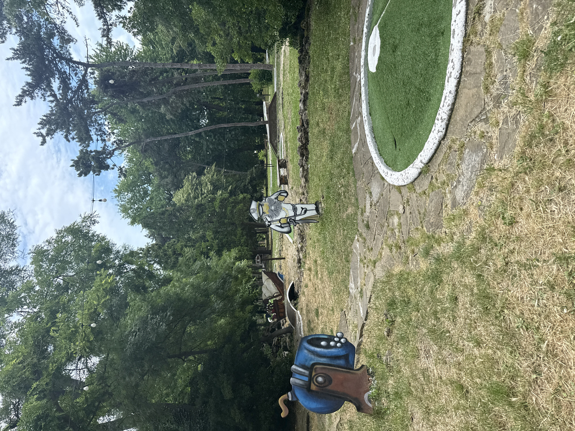 The image shows an outdoor mini-golf course. In the foreground, on the left, is a large, cartoonish blue cannon with cannonballs. To the right, a circular putting green is visible. In the mid-ground, a cardboard cutout of a cartoonish figure dressed as a captain or admiral stands near a miniature golf obstacle shaped like a pirate ship. Further in the background, more trees and what appears to be other mini-golf holes can be seen. String lights are suspended between trees overhead. The ground is a mix of grass, dry vegetation, and stone pathways. The scene is set in a park-like environment with mature trees.