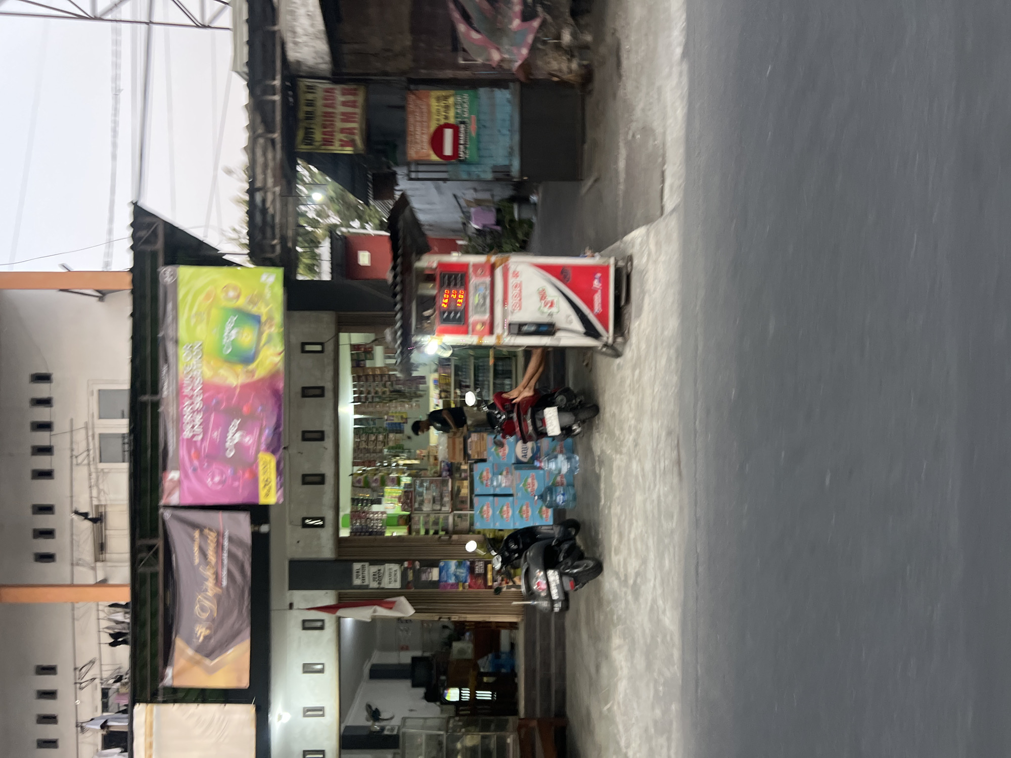 The image shows a street scene in Sendangadi, Indonesia, featuring a small convenience store or minimarket. The store has a facade with multiple windows and displays of various products, including bottled water stacked outside. Two motorcycles are parked in front of the store. A red and white gas pump stands to the right of the entrance. Banners are displayed above the storefront, one featuring a golden emblem and text, and another with graphics of fruit and text. An Indonesian flag is visible hanging near the entrance. A person is standing inside the store, and another person is sitting outside near the gas pump. The overall environment appears to be a commercial area with buildings and a paved street in the foreground.