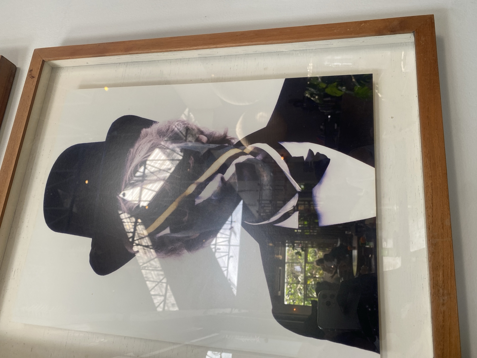 A framed portrait photograph is displayed on a white wall, possibly in the city of Mengwi, Indonesia. The photograph depicts a person from the chest up, wearing a black top hat and a black suit with a white shirt and a black bow tie. The subject's face is partially obscured by dark shadows and some colorful streaks across the image. There are reflections on the glass covering the photograph, showing an interior space with tables, chairs, and greenery, as well as a ceiling structure. The frame itself is made of wood with a distressed white matting.