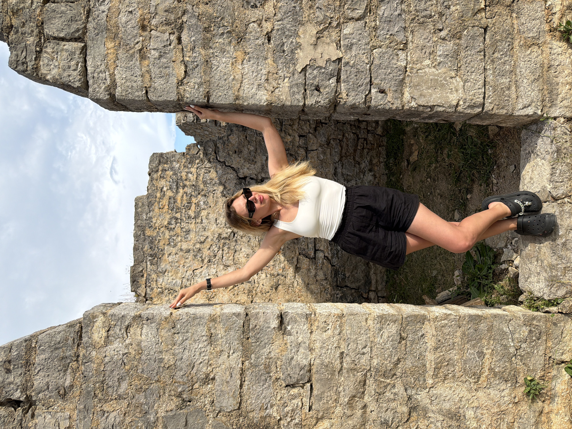 A young, light-skinned woman in a white tank top, black shorts, and black Crocs is positioned horizontally between two stone walls. She wears sunglasses and a black watch on her left wrist. Her blonde hair is straight and reaches her shoulders. Her arms are extended, each hand resting on the opposing stone wall, seemingly supporting her weight. The stone walls are gray and appear to be part of an old structure, possibly ruins, with visible cracks and uneven surfaces. In the background, beyond the structure, a blue sky with scattered white clouds is visible. There is sparse vegetation at her feet, and the location is Budva, Montenegro.