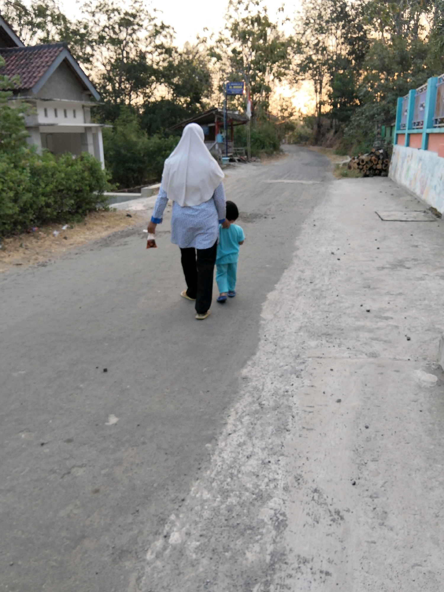In Jenangan, Indonesia, two individuals, a woman and a child, walk away from the camera down a paved road. The woman, seen from behind, wears a white hijab, a light blue and white patterned long-sleeved shirt, and black pants. She holds a clear plastic bag containing dark red items in her right hand. The child, also seen from behind, wears a light blue outfit, including pants and a long-sleeved shirt, and blue footwear. They walk on the right side of the road. The environment is rural, with trees and vegetation lining the road. A small building with a red roof is visible on the left. The sun appears to be setting, casting a warm glow on the trees in the background. The road itself is weathered and shows signs of wear.