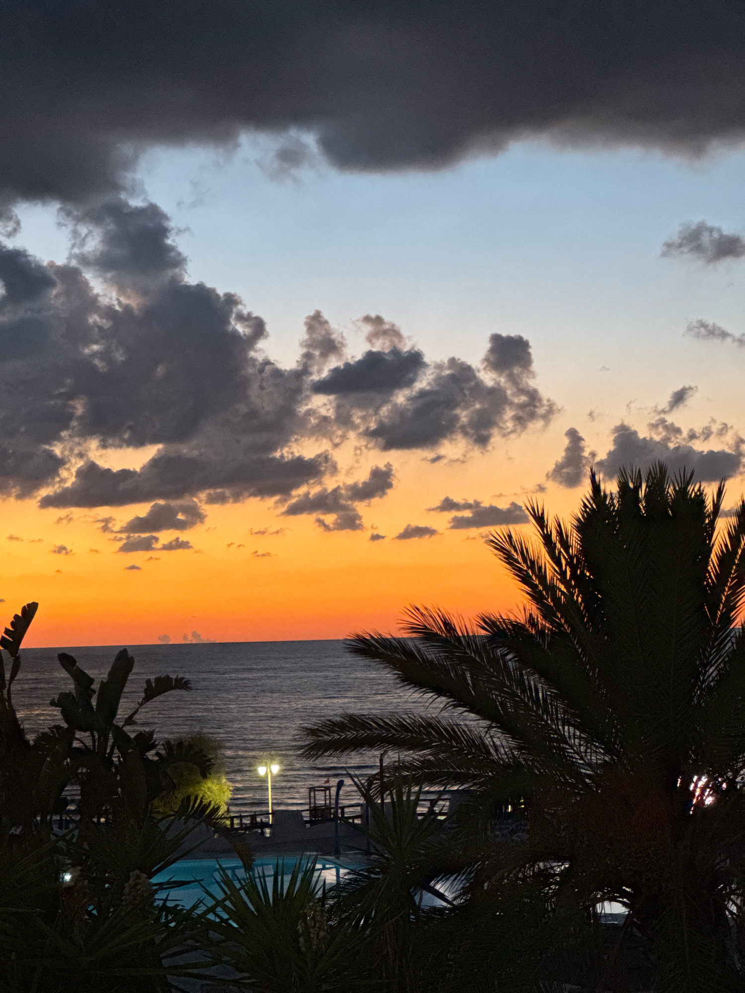 The image depicts a sunset over the sea, viewed from a vantage point with lush vegetation in the foreground. The sky is a gradient of colors, transitioning from deep gray clouds at the top to bright orange and yellow hues near the horizon. The sea is dark and appears calm. Silhouetted palm trees and other tropical plants frame the scene, obscuring parts of the view. In the middle ground, a swimming pool is visible, and a lamppost illuminates an area adjacent to it. The location appears to be a coastal resort or a residential area overlooking the Mediterranean Sea in Isola delle Femmine, Italy, given the tropical foliage and the presence of a swimming pool, suggestive of a leisure facility or private residence. There are no people or animals visible in the image. The overall atmosphere is serene and peaceful, characteristic of a twilight setting.