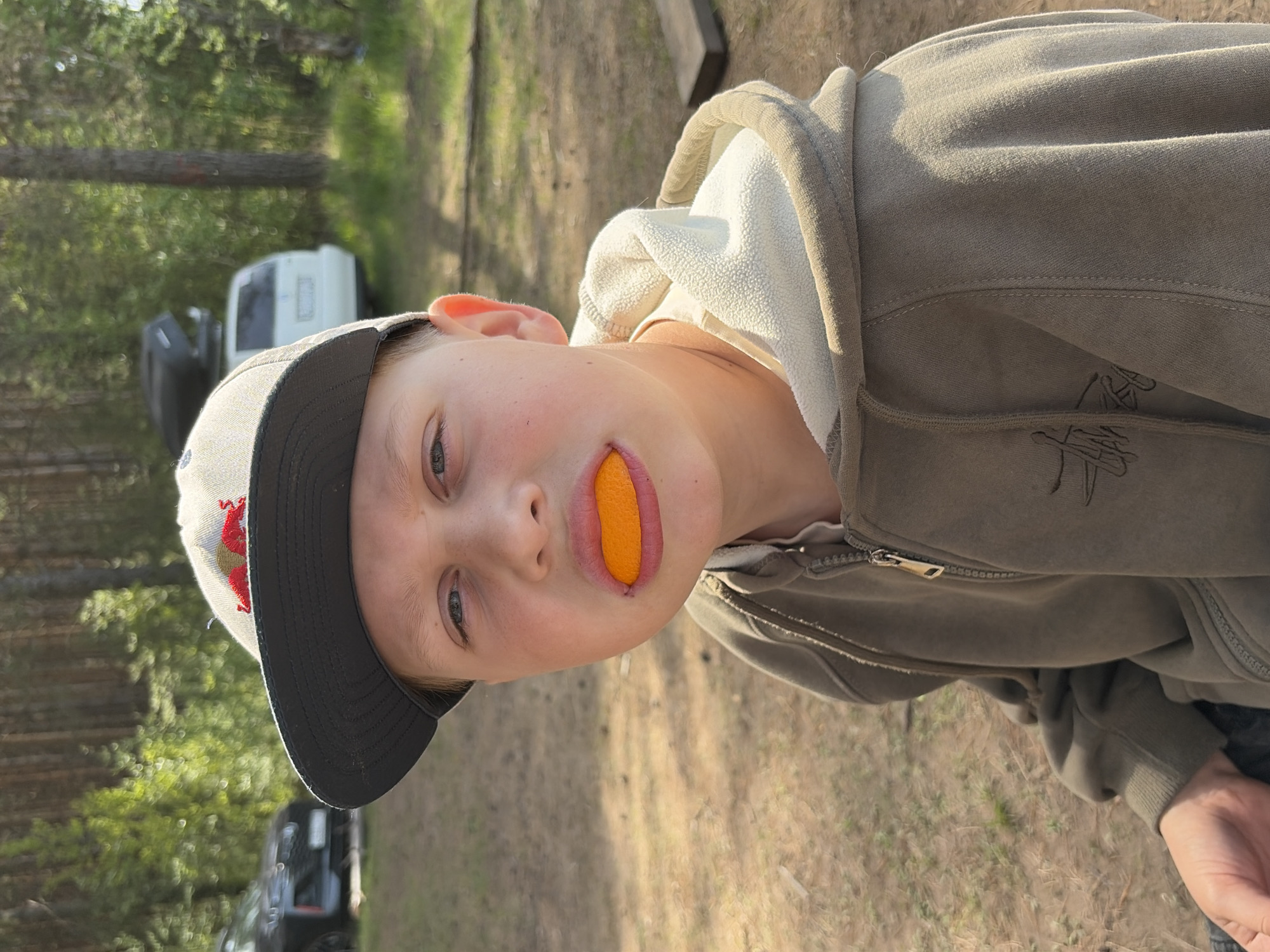 A fair-skinned, young person with blue eyes and a baseball cap is holding a piece of orange in their mouth. They are wearing a zip-up hooded sweatshirt. The location appears to be outdoors in a wooded area, with trees and vehicles visible in the background. The picture may have been taken in Забегаево, Russia.