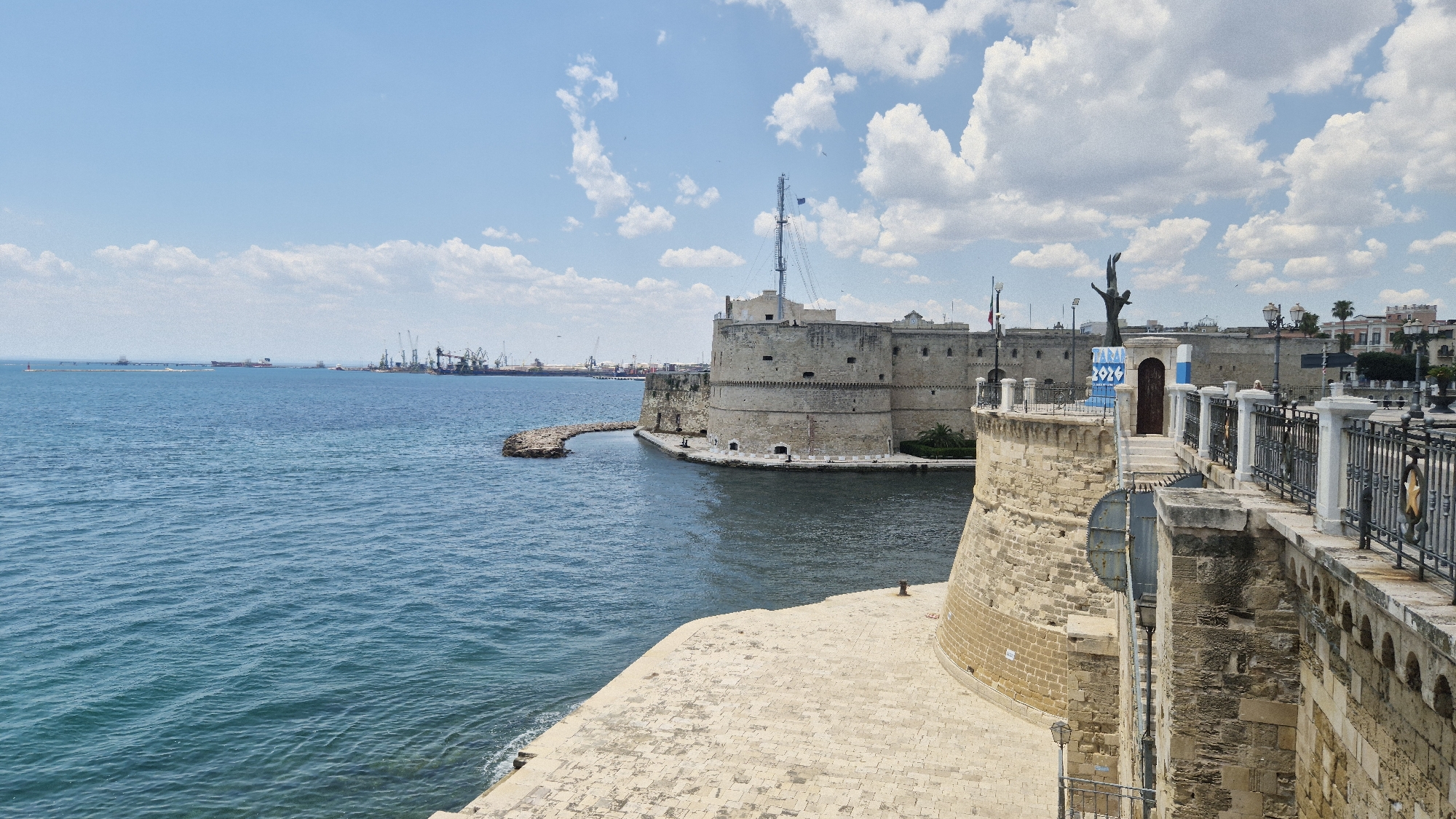 The image shows a panoramic view of the Aragonese Castle in Taranto, Italy, on a clear day with scattered clouds. The castle, a prominent historical landmark, stands on a peninsula overlooking the Ionian Sea. In the foreground, a stone promenade with a decorative metal railing leads towards the castle. A statue of a diver is visible on the promenade, with a banner that reads "TARANTO 2024". The sea is a deep blue, with gentle waves. In the background, a busy port with cranes and ships can be seen, suggesting Taranto's significance as a maritime city. The sky is a bright blue, with fluffy white clouds adding depth to the scene. The architecture of the castle is robust and ancient, characterized by its thick stone walls and battlements, indicative of its defensive purpose throughout history. The overall scene is one of historical grandeur meeting a vibrant, active waterfront.