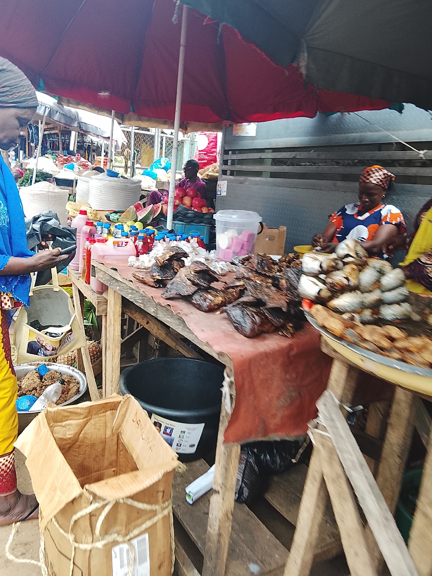 The image depicts an outdoor market scene in Akwanga, Nigeria. In the foreground, a person wearing a blue garment and a patterned headscarf is holding a dark object and a beige tote bag. To their left, a portion of a cardboard box is visible, tied with rope.
The central part of the image features a wooden stall laden with various food items. There are piles of fried fish, some whole and some in pieces, as well as what appears to be a platter of smaller fried fish or seafood in the foreground. Behind the fish, a collection of bottled drinks and containers of liquids in shades of pink and red are arranged on the stall. Further back, a woman wearing a patterned headscarf and a colorful, patterned top is seated behind the stall, actively engaged with the items in front of her. She is positioned near a plastic container holding what looks like soap bars or similar items.
In the background, other market stalls are visible, with displays of produce such as apples and watermelons. Large woven baskets containing grains or other dry goods are also present. A dark-colored tub is situated on the ground near the stall. The market is covered by a large, dark-colored umbrella or tarp, providing shade. The overall environment suggests a bustling commercial area where food and other goods are being sold.