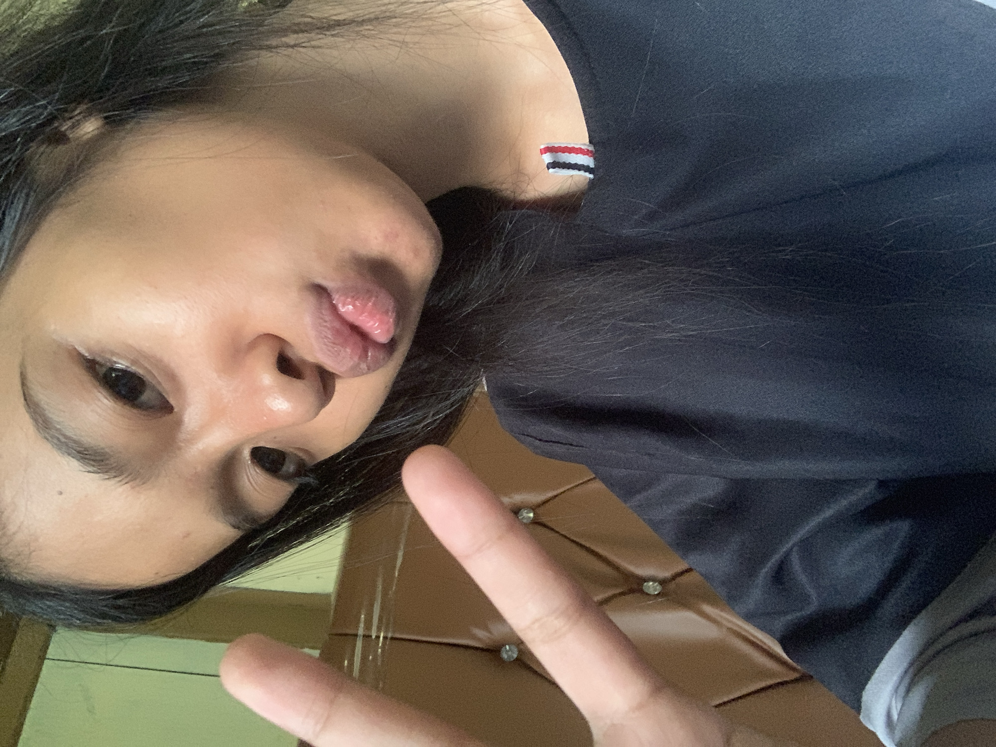 A young woman with dark hair is taking a selfie indoors, making a peace sign with her left hand and puckering her lips. She is wearing a black t-shirt with a small, striped emblem on the collar. Behind her, a brown upholstered surface with diamond-shaped stitching and clear embellishments is visible. The lighting suggests an indoor environment. The scene is likely from a residential setting in Sungai Meriam, Indonesia.