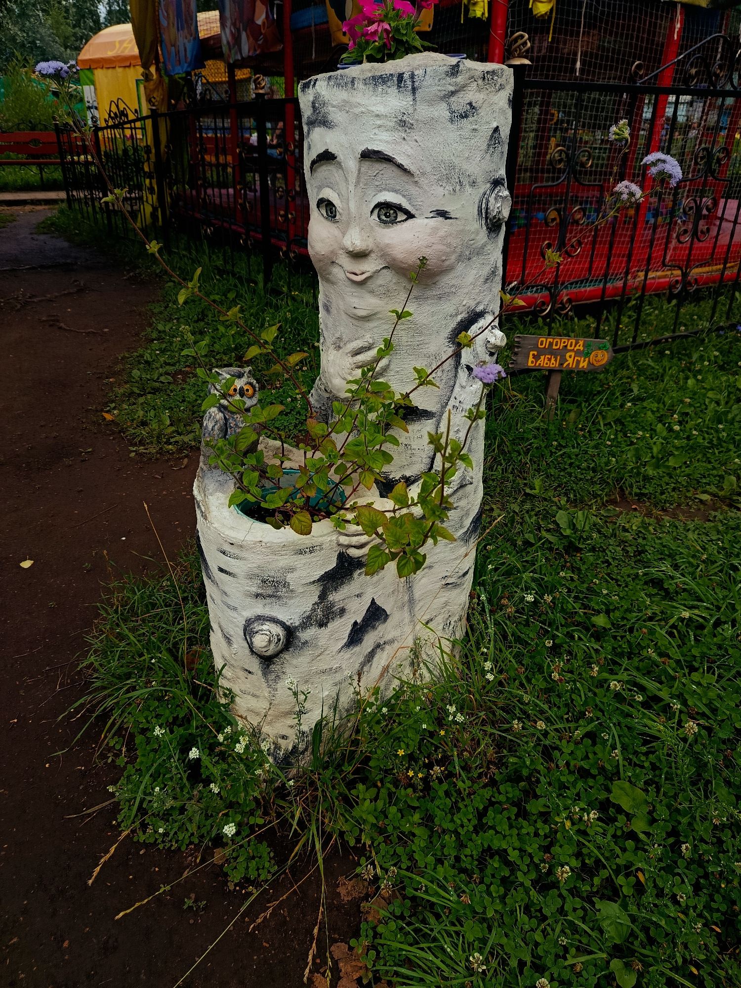 A whimsical, white-painted log sculpture with a face carved into it sits in a grassy area in Chaikovsky, Russia. The sculpture resembles a birch tree trunk with black markings and a friendly face with large eyes and rosy cheeks. A small owl figurine is perched on the sculpture to the left of the face. Green plants grow out of a planter integrated into the top of the sculpture. In the background, a black metal fence encloses a red and yellow amusement ride. A small wooden sign with Cyrillic text and a smiley face is visible near the fence. The ground around the sculpture is a mix of dirt and lush green grass dotted with small white flowers. The overall setting appears to be an outdoor recreational area or park.