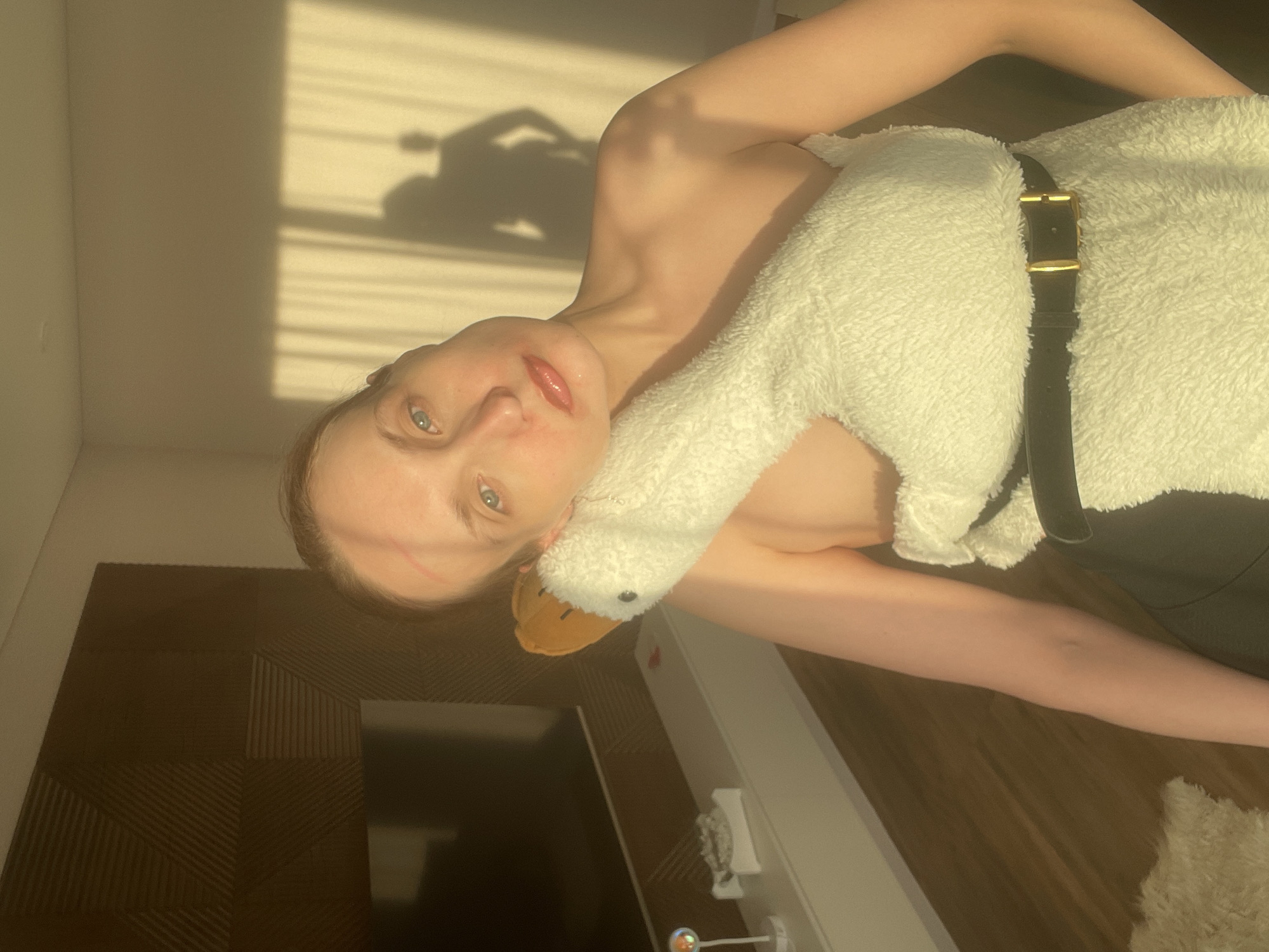 A young woman is captured in a close-up, half-length shot, lying on her side and facing the camera with a neutral expression. She has light blue eyes and shiny, glossed lips. Her bare shoulders and arm are visible, and she is holding a large, white, fluffy stuffed animal that resembles a goose or swan, which is cinched with a black belt featuring a gold buckle.
The background is bathed in warm, directional sunlight, casting distinct, elongated shadows on a light-colored wall. One shadow clearly depicts a person holding a camera or phone, suggesting a selfie or self-portrait. A dark, textured surface, possibly wood flooring or furniture, occupies the lower and left portions of the frame. A white piece of furniture with a small light fixture or decorative item is also visible. The strong sunlight indicates it is daytime. There is no visible text in the image. The location context provided is Подшивалово, Russia.