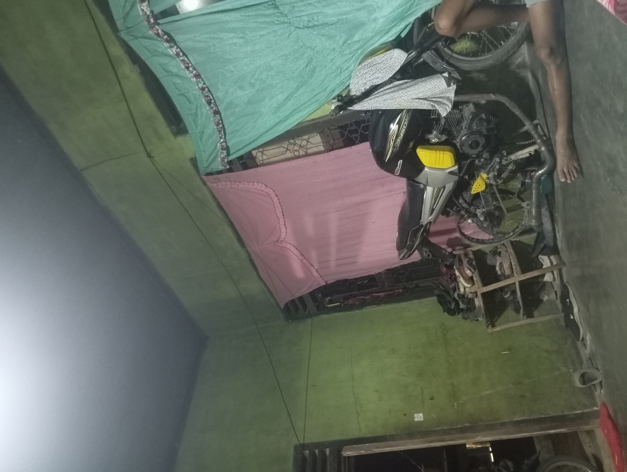 In a dimly lit room in Rejosari, Indonesia, a black and silver motorcycle with yellow accents is parked. A light-colored garment is draped over the motorcycle's handlebars. To the left of the motorcycle, a pink fabric is hanging, and to the right, a teal fabric hangs. In the foreground, a person's leg and foot are visible, suggesting they are sitting or lying down. To the left, a wooden shoe rack is filled with various pairs of shoes. The walls of the room are painted green, and the floor appears to be concrete. The overall impression is one of a lived-in space, possibly a home or garage, with everyday objects present.