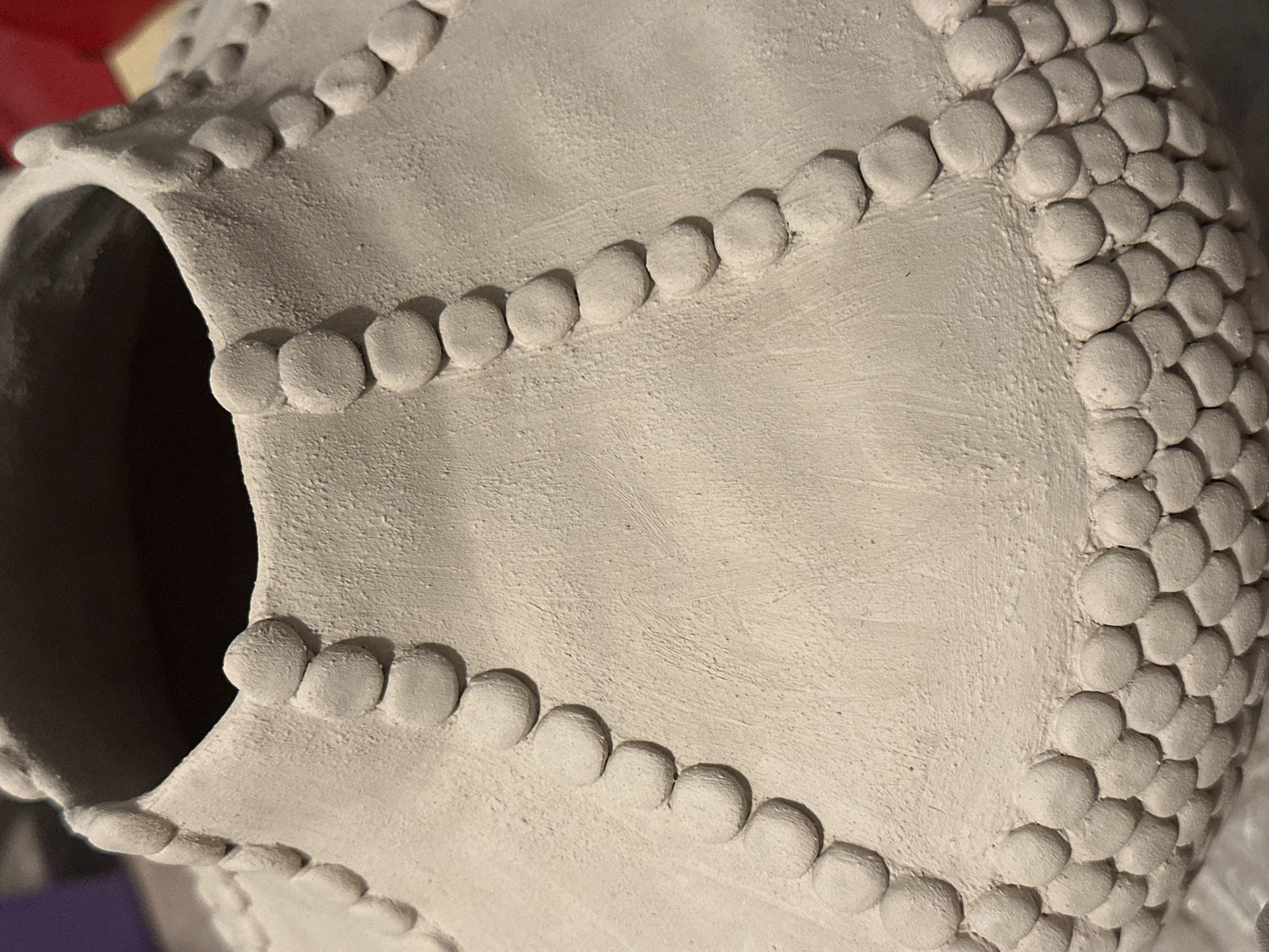 The image shows a close-up, eye-level view of a light-colored, unglazed ceramic vase. The vase has a textured surface and is adorned with rows of small, rounded clay appliques that resemble beads or scales. These appliques are arranged in decorative patterns, including lines that fan out from the neck of the vase and a dense, overlapping pattern around its base. The lighting creates subtle shadows that highlight the three-dimensional texture of the appliques. The background is out of focus, with hints of darker and lighter colors, suggesting an indoor setting. There is no visible indication of the city of Nevinnomyssk, Russia.