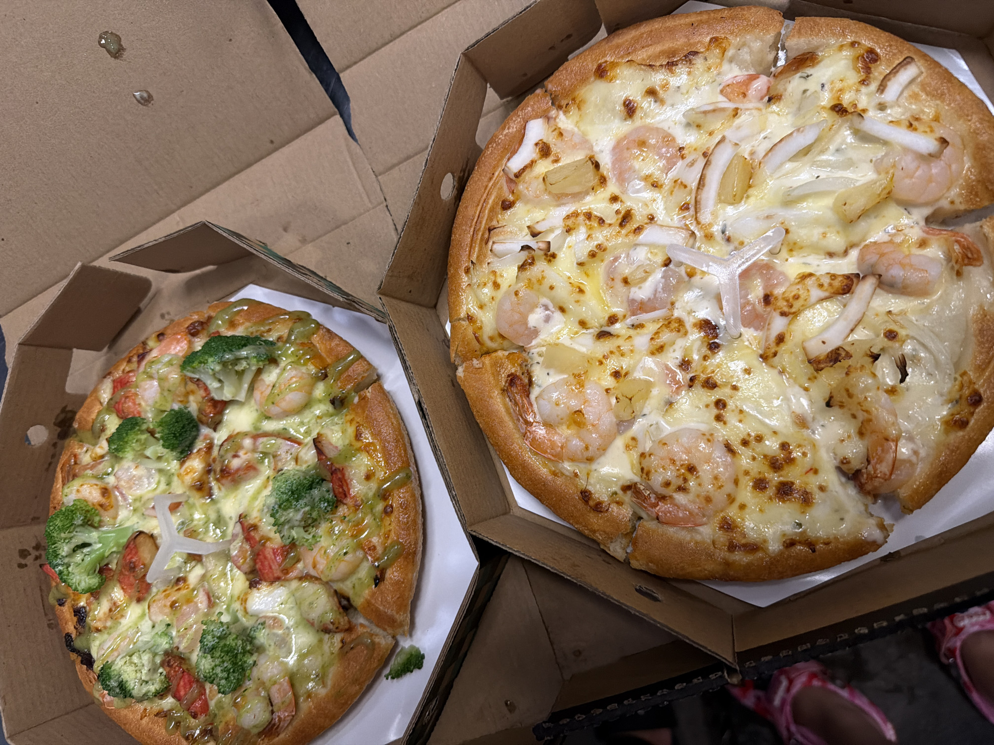 The image shows two pizzas in open cardboard boxes, likely for take-out. One pizza, on the left, has broccoli, crab sticks, and shrimp as toppings, with a green sauce drizzled over it. The other pizza, on the right, has shrimp, squid, and pineapple as toppings. A plastic pizza saver is placed in the center of each pizza. The scene is illuminated by indoor lighting. The background is indistinct but suggests an interior setting. Based on the presence of Vietnamese text in the image metadata, it is likely located in Đà Nẵng, Vietnam.
