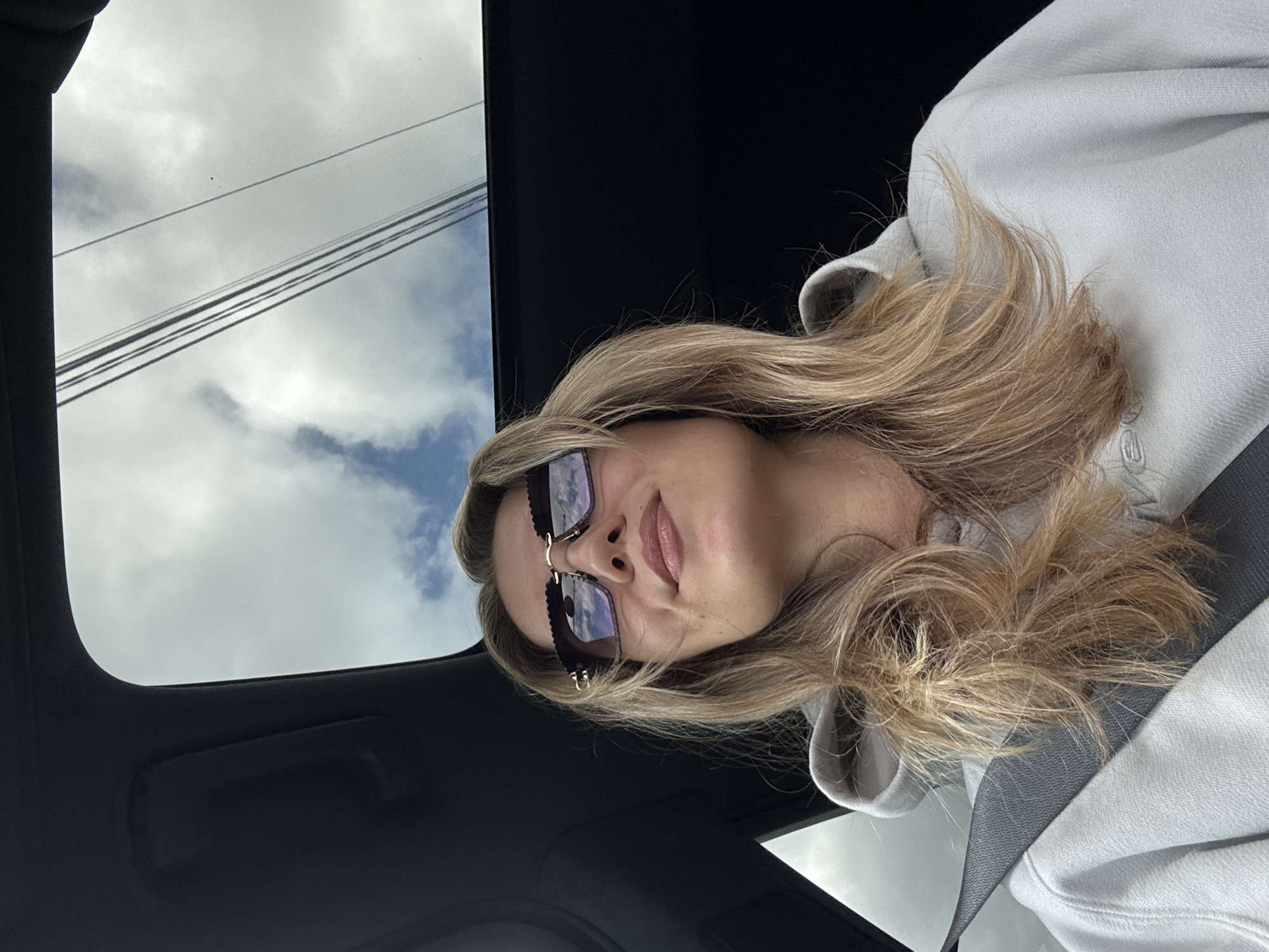 The image, oriented vertically and rotated 90 degrees counter-clockwise, captures a person with long, wavy blonde hair looking upwards from the interior of a vehicle. The individual, positioned on the right side of the frame, wears square-framed sunglasses with dark lenses and distinctive gold-toned, wavy-edged frames. Their lips are slightly parted. They are dressed in a light gray hooded sweatshirt, featuring a faint, stylized "M" logo visible near the collar on the right side of the fabric. A dark gray seatbelt is fastened across their chest.
The setting is the interior of a car, evident from the dark headliner and trim. Through an open sunroof, a bright, partly cloudy sky is visible, showing both white clouds and patches of blue sky. Several utility wires or power lines are stretched across the sky in the upper left, viewed through the sunroof. The reflections of the sky and clouds are clearly visible in the person's sunglass lenses. The scene depicts a daytime moment, likely a selfie taken while in transit.