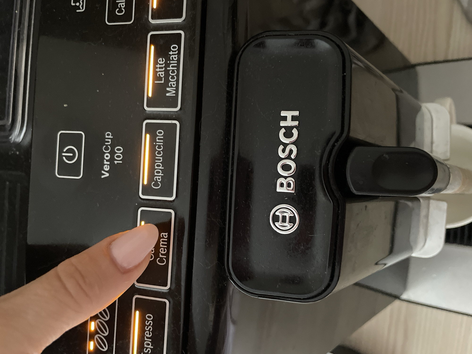 A person's index finger, with a light pink nail, is pressing the "Crema" button on a black Bosch coffee machine. The button is illuminated with an orange light. Other illuminated buttons include "Espresso" and "Cappuccino" to the left, and "Latte Macchiato" to the right. Above the buttons, the text "VeroCup 100" is visible, along with a power symbol. The Bosch logo is prominently displayed on the front of the machine. The background shows a light-colored wooden surface and a white cup, suggesting the coffee machine is in a home or office setting.