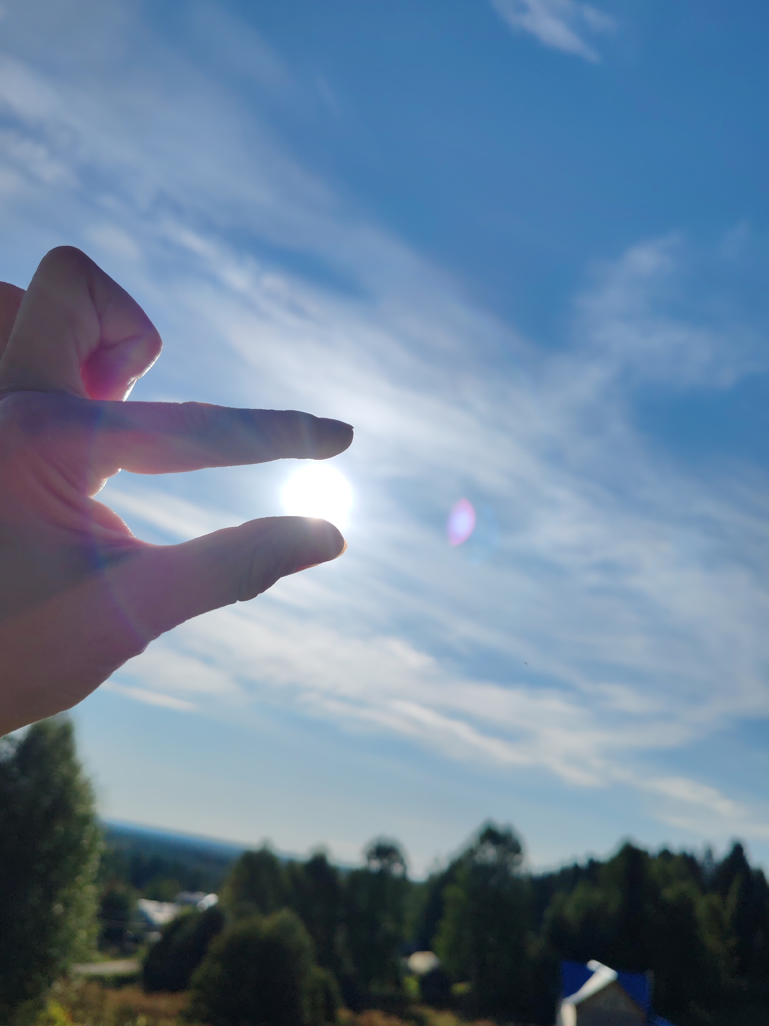 A person's hand is positioned in the foreground, silhouetted against a bright sun and a blue sky with scattered white clouds. The thumb and index finger of the hand are curved towards each other, framing the sun. The sun appears as a bright, slightly diffused orb with lens flare. The background shows a blurred landscape of trees and some buildings, suggesting an outdoor scene. The image appears to be taken in Чухлэм, Russia.