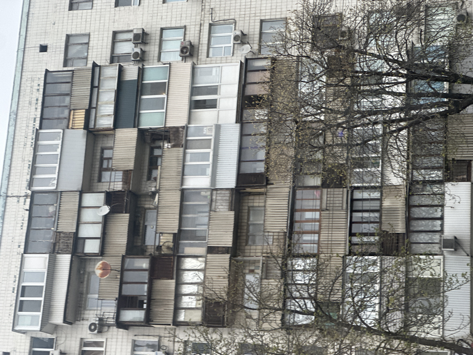 The image shows the exterior of an apartment building in Kyiv, Ukraine. The facade is characterized by a chaotic and mismatched collection of enclosed balconies and window modifications. Various types of materials, including corrugated metal and different styles of window frames, are used to enclose the balconies. Several satellite dishes are affixed to the building's facade. There is bare tree branches partially obstructing the view. The building is primarily white or light gray.
