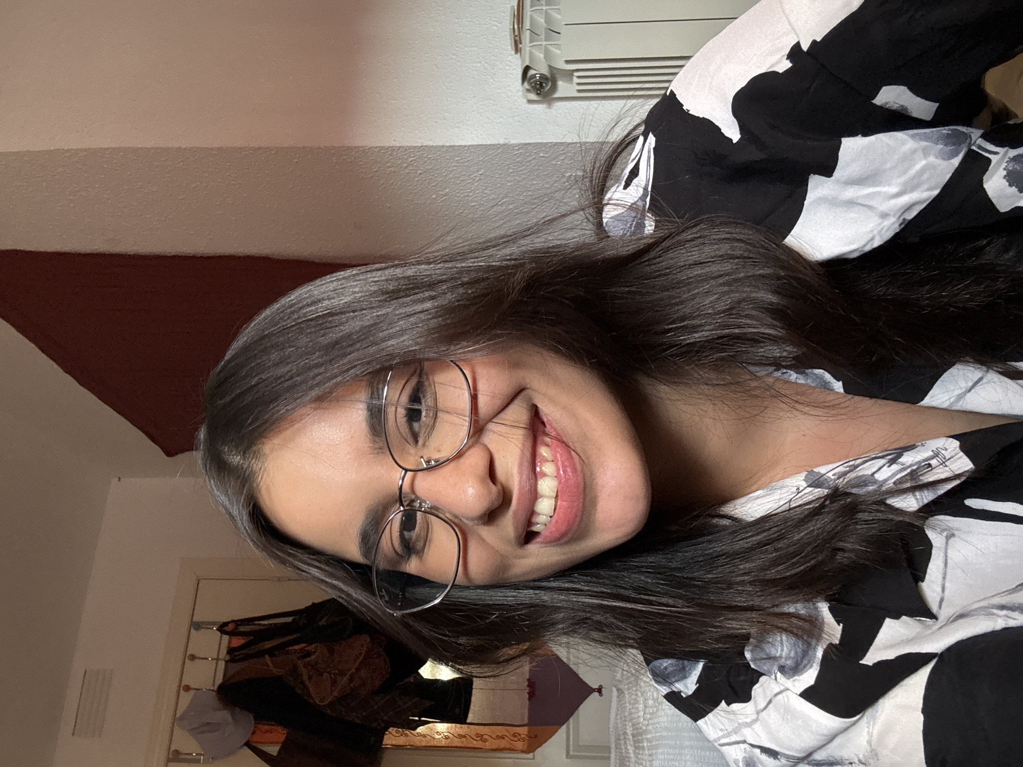 A young woman with long, dark hair and glasses is smiling at the camera in a close-up portrait. She is wearing a black and white patterned shirt. The background is slightly blurred, showing a wall with a dark red fabric hanging on it, and a radiator. To the left, there's a door with several items hanging on hooks. The scene appears to be indoors, possibly in a bedroom or living space. The lighting is soft and natural.