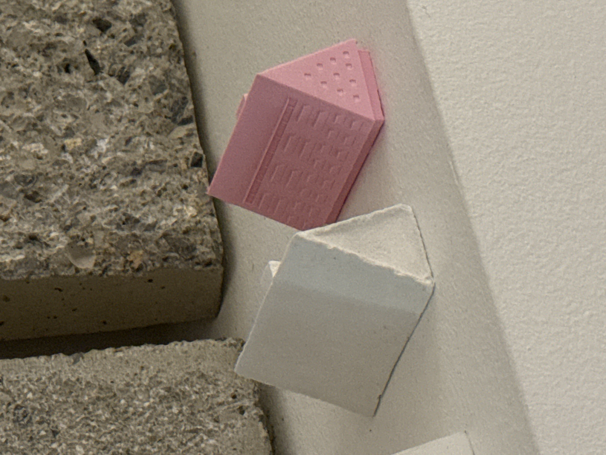 The image depicts two small, stylized building models. One is pink and appears to represent a skyscraper with visible windows and a roofline featuring small, regularly spaced indentations. The second model is white and also suggests a building, though with fewer visible details. These models are positioned on or against a textured, neutral-colored surface, possibly a wall or other architectural element. Next to the models is a rough, gray stone block. While the image is indoors, the location cannot be determined with certainty, and there is no explicit information to confirm it is in Milan, Italy.