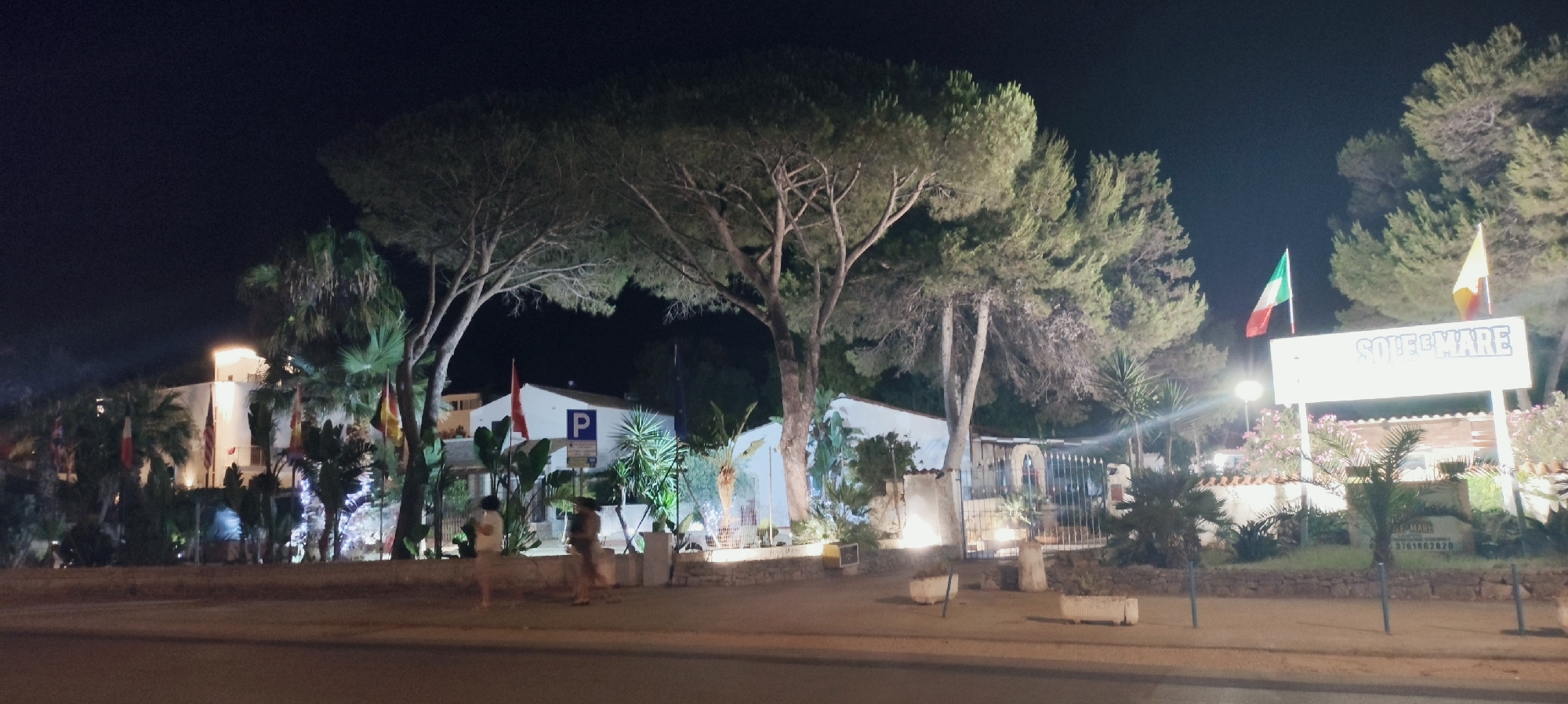 The image depicts a nighttime scene outside a building, likely a hotel or restaurant, in San Vito Lo Capo, Italy. The building is white with illuminated windows and architectural features. It is surrounded by lush greenery, including palm trees and large pine trees. Several flags are visible, including the Italian flag. A prominent sign displays "SOLE E MARE" in large blue lettering. Below this, another sign with text and a phone number is partially visible. A parking sign with a blue 'P' is also mounted on the building. In the foreground, there is a paved area with some planters and a road. Two blurry figures, appearing to be pedestrians, are visible on the paved area. The lighting suggests it is evening, with artificial lights illuminating the building and its surroundings.