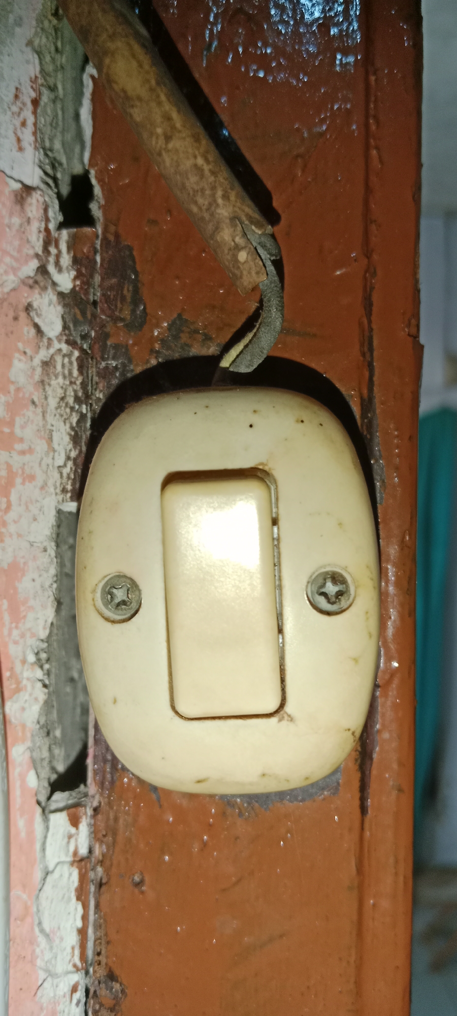 The image shows a close-up of an old, cream-colored light switch mounted on a brown, painted surface, likely a door frame or wall. A weathered, dark-colored wire protrudes from the top and connects to the switch. The wall to the left of the switch is peeling, revealing a rough, textured surface underneath. The light switch itself has two visible screws holding it in place, and a rectangular button in the center. The scene appears to be in an indoor setting, possibly an older building. The location is specified as Subang, Indonesia, suggesting this is a typical residential or older commercial structure found in that region.