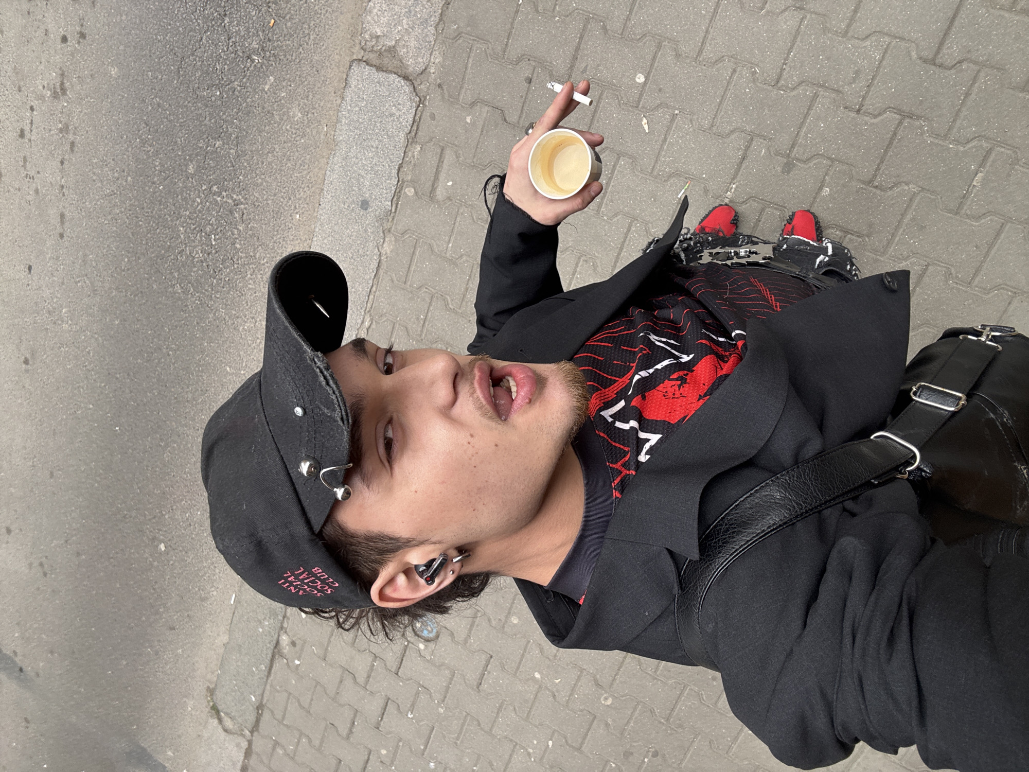 A young male individual is depicted from a high-angle, eye-level perspective, standing on a sidewalk in an urban environment identified as Sofia, Bulgaria. The subject wears a black baseball cap adorned with pink "ANTI SOCIAL SOCIAL CLUB" text and multiple silver piercings. He has dark, curly hair, a goatee, and is winking his right eye with his mouth open, looking directly at the viewer. A black wireless earbud with red accents and a silver hoop earring are visible in his left ear.
He is dressed in a black long-sleeved jacket with frayed cuffs, layered over a red and black patterned top. A black textured shoulder bag with silver hardware is worn on his left side. In his right hand, he holds a small white paper cup containing a light-brown liquid (likely coffee) and a lit cigarette between his index and middle fingers. Distressed red and black footwear is visible at the bottom of the frame. The ground beneath him is composed of light grey interlocking concrete pavers, bordered by a concrete curb separating it from a darker grey asphalt road. The scene is illuminated by diffused natural light.