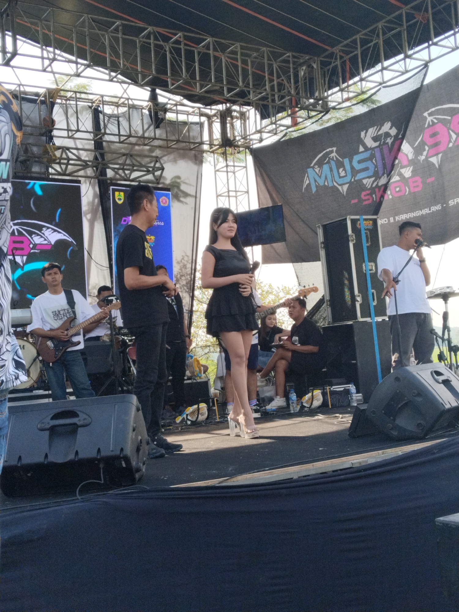 The image shows a band performing on an outdoor stage. There are several members visible, including two singers with microphones, a guitarist wearing a white t-shirt with the words “SUICIDE SILENCE”, and other instrumentalists. Behind the band, there are banners with the word “MUSIK 99 SKOB” visible on them. The banners also have the phrase “NO II KARANGMALANG - SA…” written. This location is in the city of Karanganom, Indonesia. The stage is constructed with metal scaffolding, and there are monitors and speakers visible.
