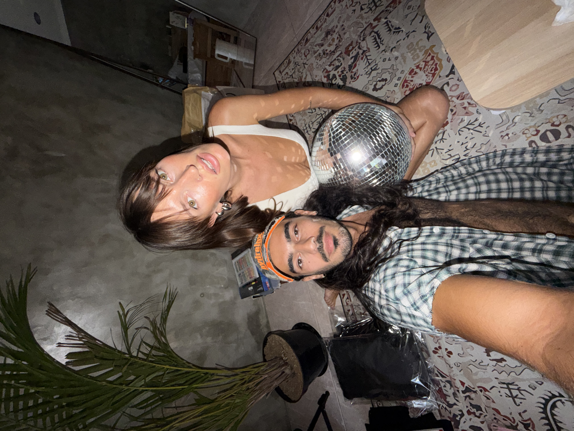 This overhead, rotated image captures two individuals lying on a floor or rug indoors, possibly in North Kuta, Indonesia.
The primary subjects are a woman and a man. The woman, with long dark hair and striking light-colored eyes, wears a white top and gazes directly at the camera. She holds a large, reflective disco ball against her chest and shoulder. The man, with dark, wavy hair and a beard, is wearing a green and white plaid shirt and an orange and camouflage patterned headband. He also looks towards the camera with a neutral expression.
The setting appears to be a casual indoor space, characterized by a detailed, patterned rug covering a significant portion of the floor, alongside a darker concrete or tiled area. A potted, palm-like plant stands in the lower left, adding a touch of greenery. In the background, hints of furniture, possibly a wooden table corner, and various packages or items suggest a lived-in environment.
The activity appears to be a relaxed pose for the camera, possibly during an informal gathering or photoshoot. The presence of the disco ball suggests a festive or playful atmosphere, perhaps indicating an evening or night-time event. The lighting is artificial, casting reflections and highlights, particularly on the disco ball and the subjects' skin. No discernible text is clearly visible in the image. The overall mood is relaxed and engaged.