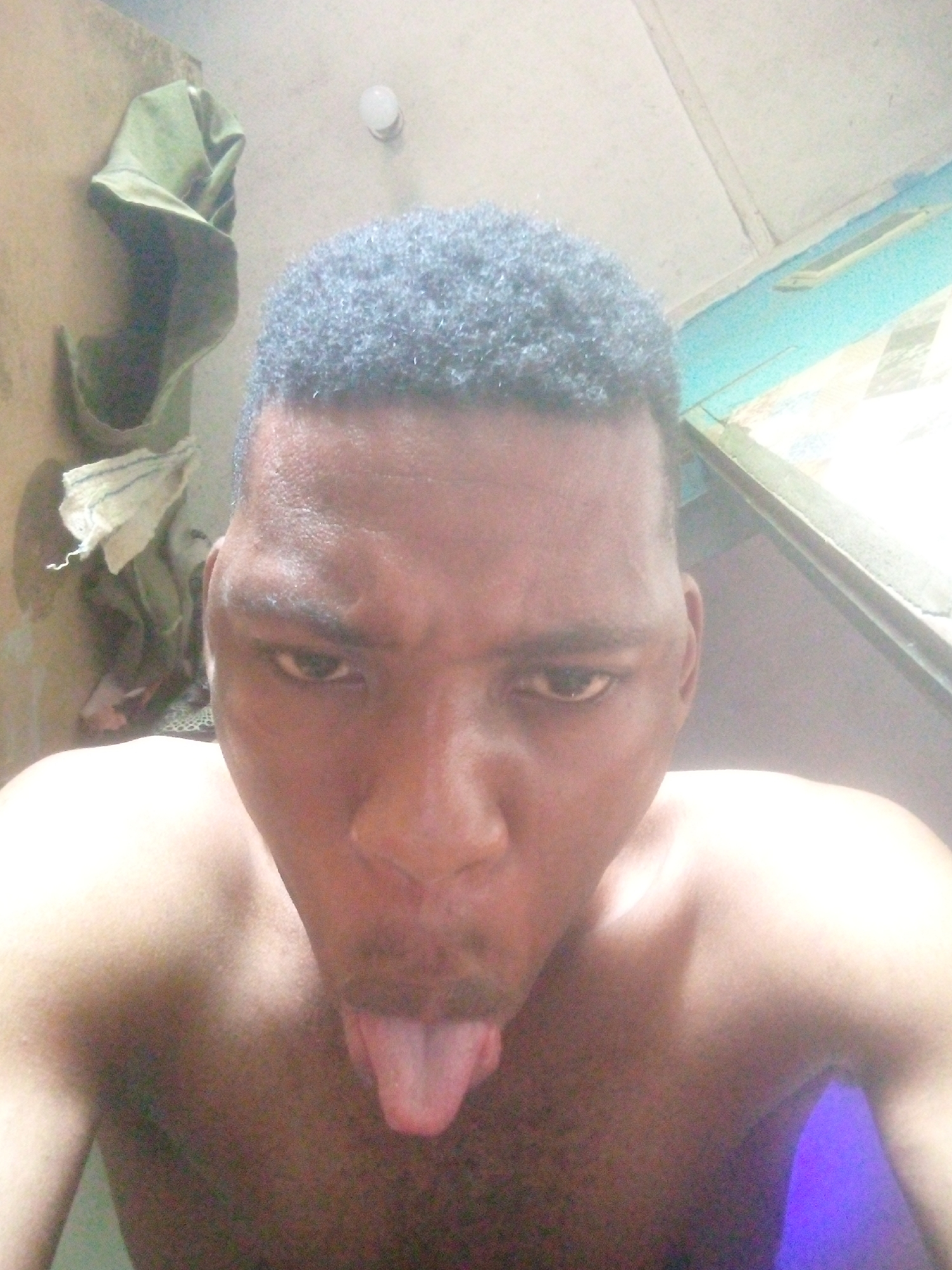 A close-up, high-angle selfie depicts a shirtless man with dark skin and short, curly black hair. He is sticking his tongue out and looking at the camera. Behind him and to the left, a green hat and a white cloth hang on a wall. Above him, a light bulb is visible on the ceiling. A light blue structure is visible in the background to the right. The location appears to be indoors. The city and state are unknown.