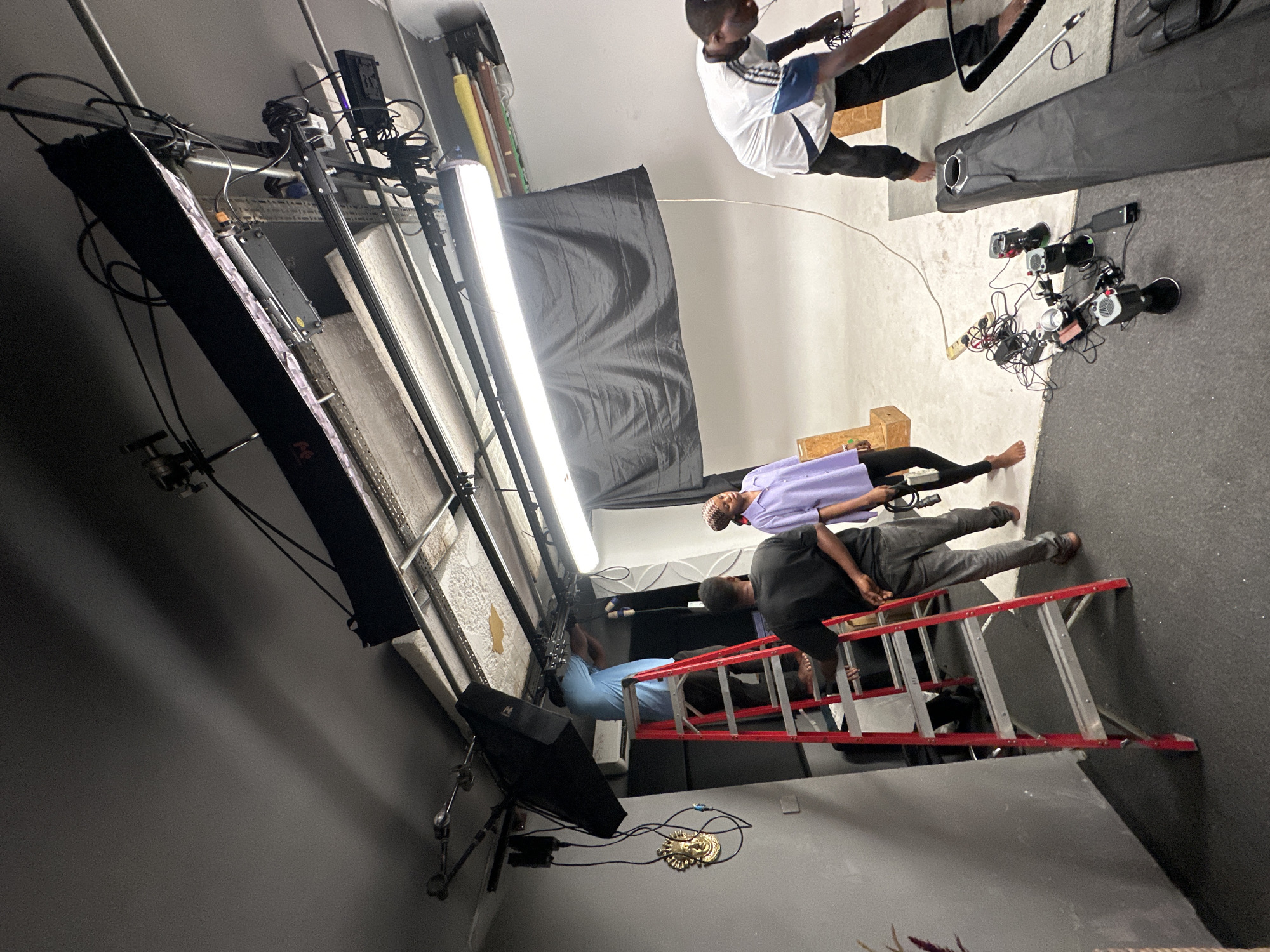 The image depicts an interior scene, likely a photography or film studio, in what appears to be Ikeja, Nigeria.
In the foreground, a red and silver ladder is positioned, with at least one person wearing a blue shirt standing on it. Above the ladder, a person in a black shirt leans over, interacting with someone wearing a light purple shirt who is lying on the floor.
To the right, a person in a white and blue shirt lies on the floor. There are photography or film lighting equipment, including boom stands, softboxes, and camera lights, strewn around the studio floor. A large black backdrop is suspended in the background. Cables are visible. The floor is partially covered in dark gray carpeting. The walls are light gray. There's what appears to be a small, golden decorative item on the floor. The overall lighting in the studio is bright.