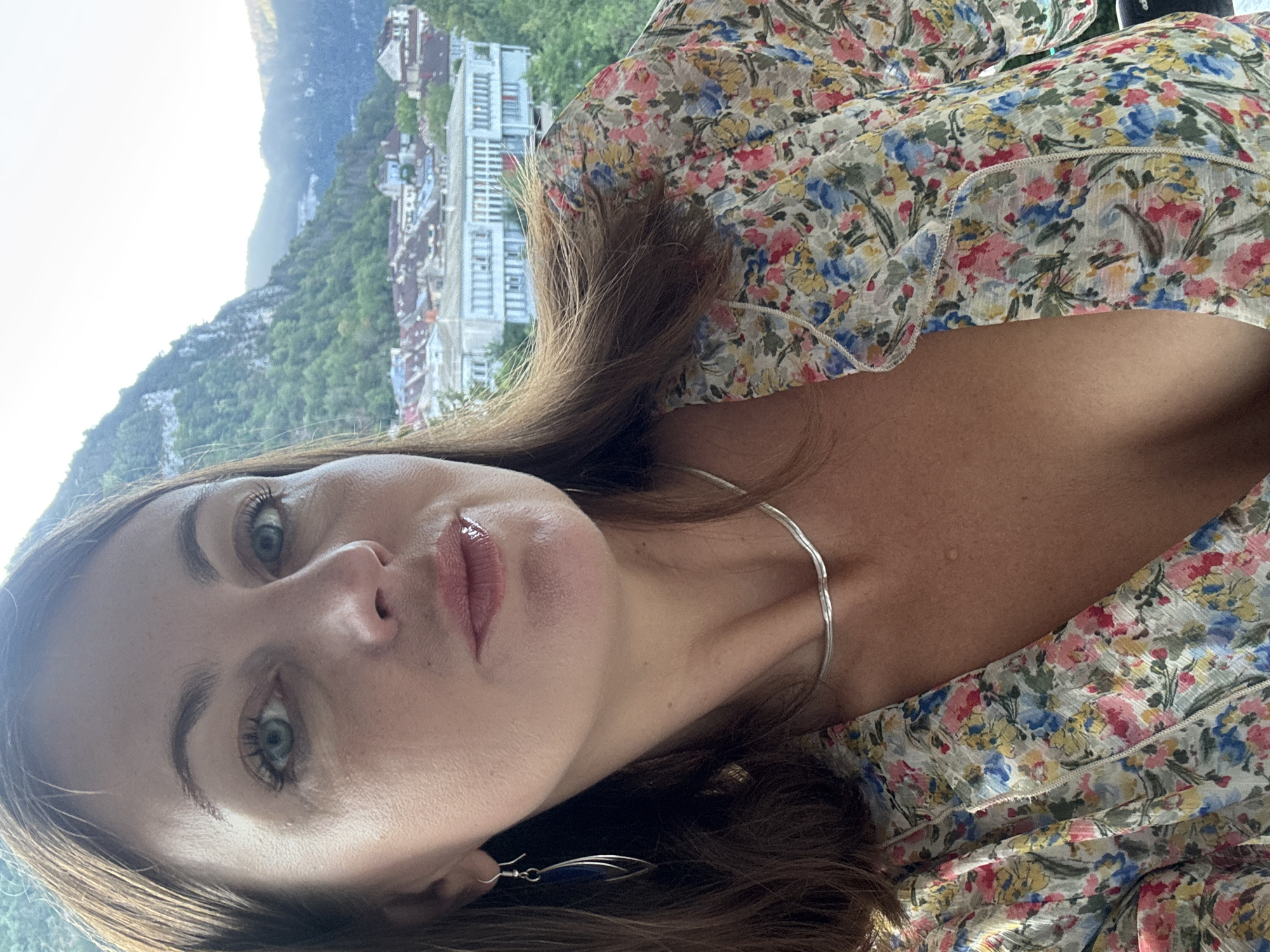 A woman with brown hair and blue eyes, wearing a floral print dress, a silver necklace, and blue earrings, poses for a selfie with a town and mountains in the background. The town, which appears to be Saint-Claude, France, is nestled in a valley. The woman's face fills the foreground of the image, her gaze directed at the camera. The floral dress is detailed with a variety of colorful flowers. The background shows buildings with red-tiled roofs and lush green mountains. The lighting suggests it is daytime.