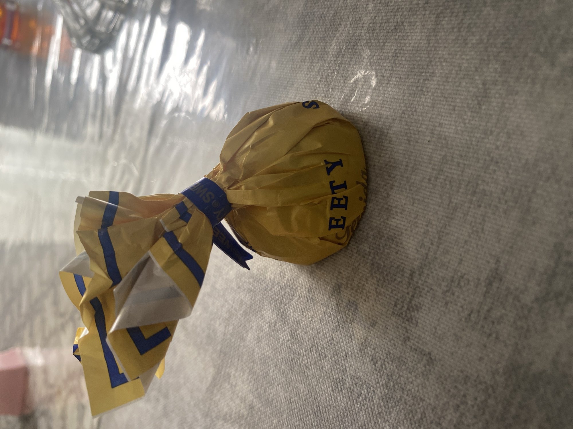 A single, unwrapped chocolate candy, wrapped in crinkled yellow foil with blue accents, rests on a gray textured surface. The foil is twisted at the top, with some of the wrapper extending upwards. The word "SWEETY" is printed in blue capital letters on the lower portion of the foil. The background is blurred, showing a shiny, reflective surface and indistinct objects. There is no indication of the city of Chontaul, Russia.