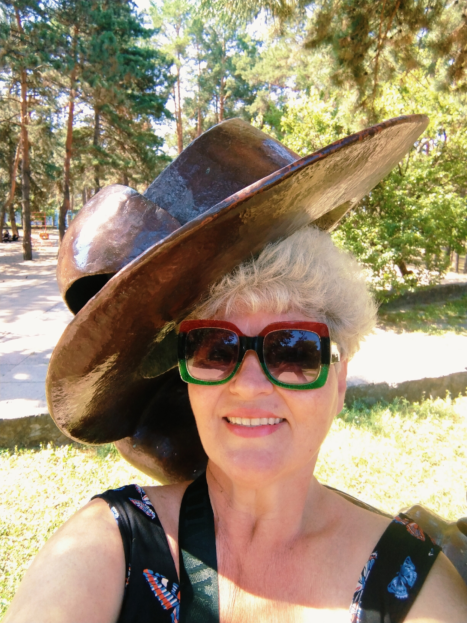 A woman with short, gray hair and a wide smile is taking a selfie outdoors. She is wearing large, rectangular sunglasses with red and green frames, and a black dress with a butterfly pattern. She is positioned behind a large, metallic sculpture of a hat, which partially obscures her head and shoulders. The background consists of trees and dappled sunlight, suggesting a park or outdoor recreational area. The presence of pine trees and the general environment are consistent with locations in Horishni Plavni, Ukraine, a city known for its industrial landscape surrounded by nature.