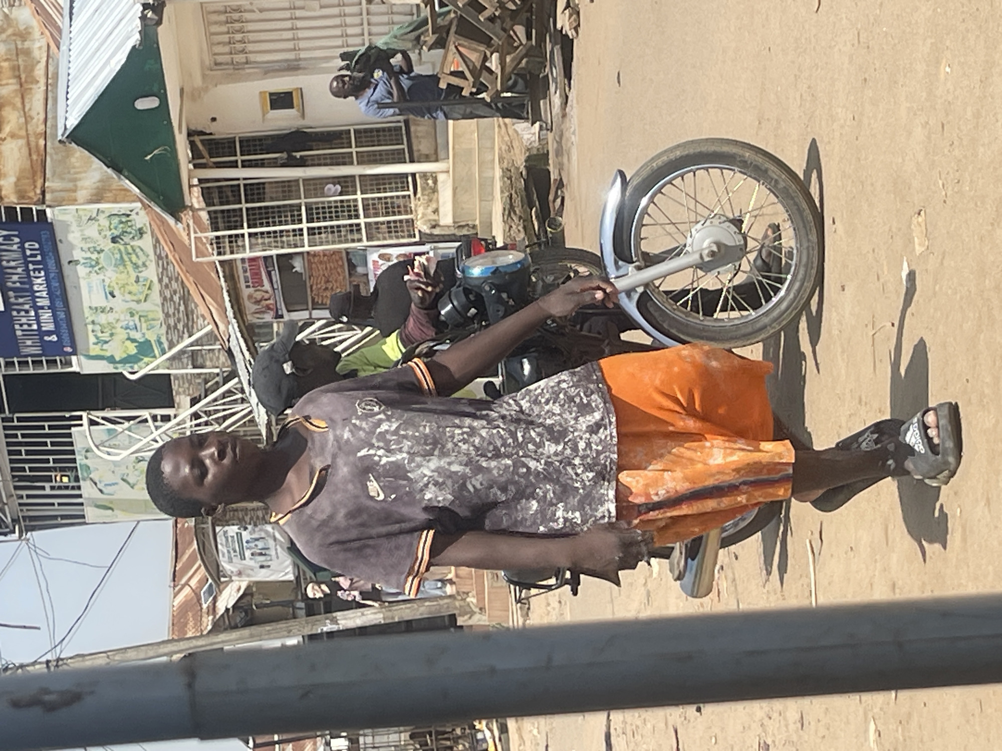 In an outdoor, dusty street scene in Abuja, Nigeria, a young Black male, appearing to be in his late teens or early twenties, walks towards the right of the frame. He wears a dark gray t-shirt with a mottled pattern and orange trim around the collar and sleeves, and orange shorts with a vertical dark stripe and a red stripe down the side. He is also wearing dark slide sandals. His left hand is partially visible and appears to be holding onto something.
Behind him, a motorcycle is parked, with its front wheel and fender prominently in the foreground on the right. Another Black male, wearing a black cap and a green shirt, is seated on the motorcycle. Further in the background, on the right, another Black male in a blue shirt and dark pants is standing near a pile of wooden structures.
The environment features buildings with corrugated metal roofs and some with green accents. A sign for "WHITEHEART PHARMACY & MINI-MARKET LTD" is visible on one of the buildings. The ground is covered in dirt and sand, with some debris scattered around. The lighting suggests it is daytime with bright sunlight.