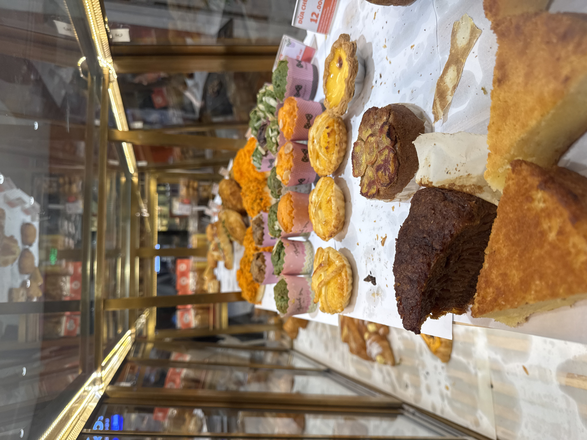 The image shows a display of baked goods in what appears to be a bakery or patisserie. The display is illuminated by warm lighting, suggesting an indoor setting, possibly a shop window or interior display case. Various pastries are arranged on a white surface, some in paper cups, and others as slices or whole cakes. A price tag is visible in the foreground, showing "12.000" and "DUA CHE". This suggests the location is in Da Lat, Vietnam, as "Dua Che" can refer to Vietnamese dessert soup or other sweet dishes, and the currency is Vietnamese Dong. There are no people clearly visible in the image, but the reflection in the glass display case suggests a shop environment. The baked goods include muffins with green and pink wrappers, small tarts with fruit toppings, slices of cake, and what looks like a loaf cake topped with banana slices. The overall impression is one of a well-stocked bakery offering a variety of sweet treats.
