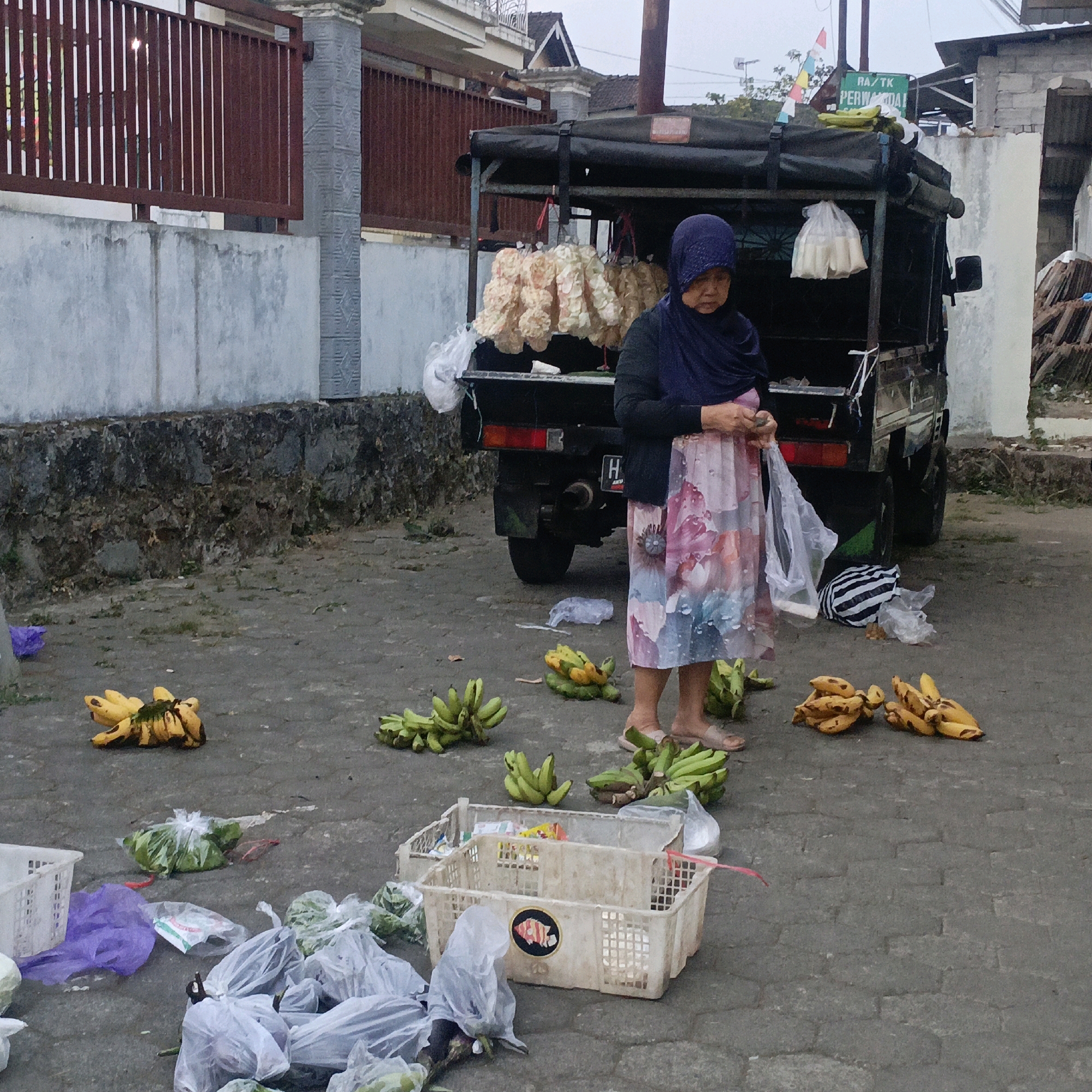 A woman wearing a blue headscarf and a floral dress stands next to a small truck, holding a plastic bag. The truck is loaded with goods, including hanging bunches of light-colored food items. Bananas are scattered on the ground around the truck, some in bunches and some individually. Several plastic bags and baskets containing produce are also on the ground. The scene appears to be a street market. A sign on the truck reads "RA/TK PERWIDAI," which indicates the location is likely a kindergarten or early childhood education center in Salatiga, Indonesia, suggesting the truck may be part of a school or community event.