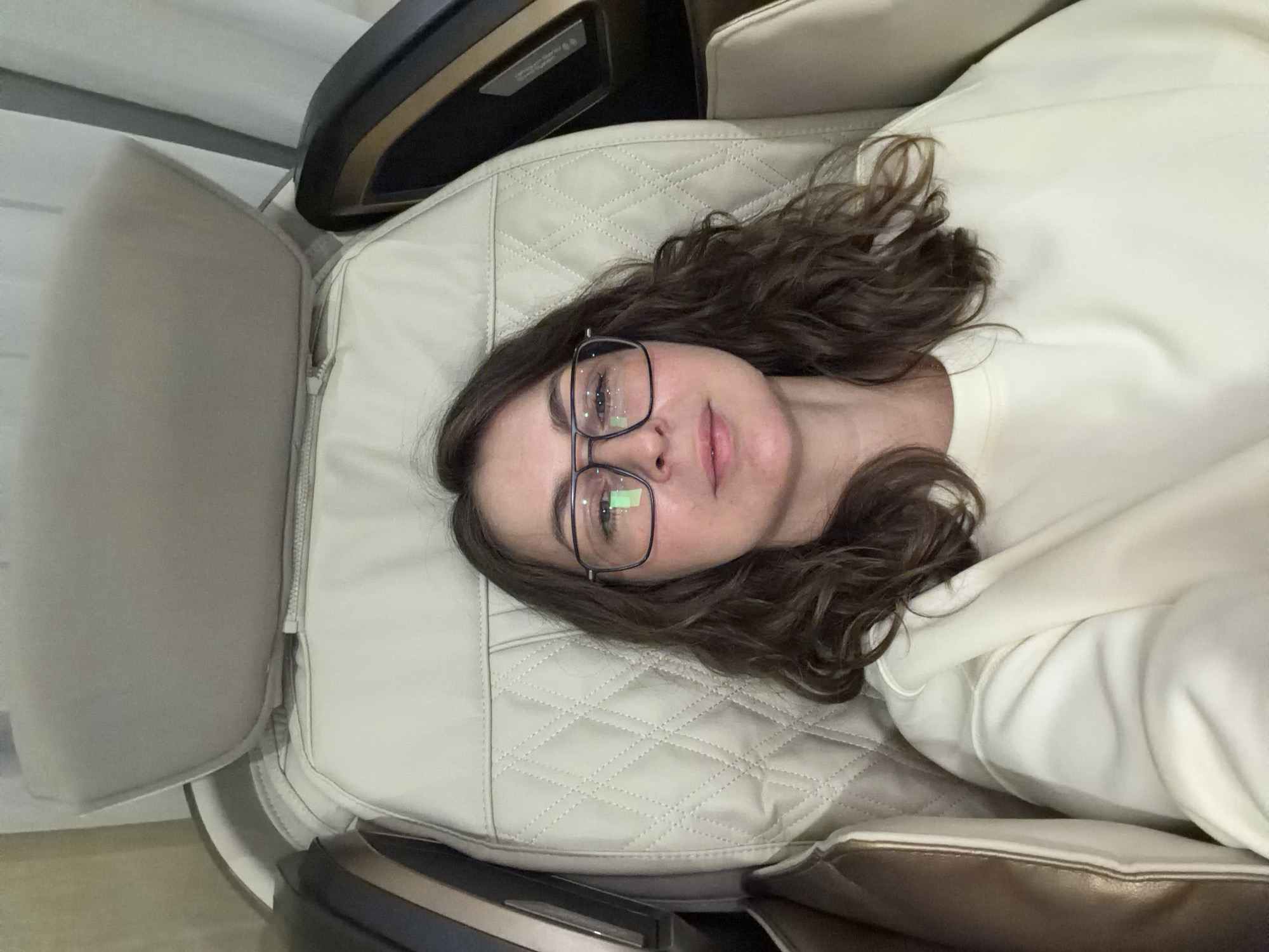 A medium shot shows a woman with brown, shoulder-length curly hair and glasses lying on a light beige leather recliner. She wears a light cream-colored sweater. The recliner has quilted detailing. The top and bottom of the recliner are visible, with dark brown and gold trim. The background is a white wall. The photograph appears to be taken from an overhead perspective. No location information for the city of Izhevsk, Russia is determinable from the image.