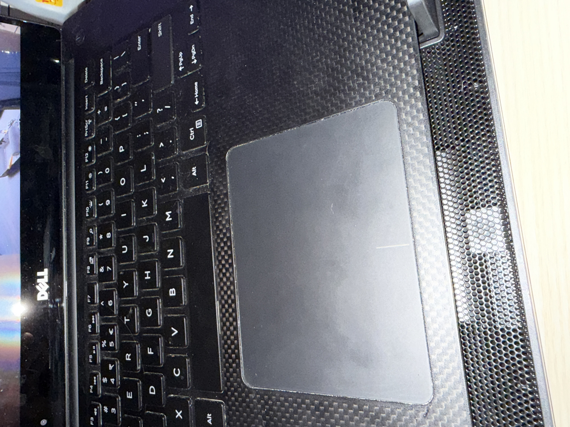 The image shows a close-up view of a black Dell laptop keyboard and trackpad. The keyboard has standard QWERTY layout with function keys at the top. The trackpad is large and centrally located below the keyboard. The laptop casing appears to be made of a textured carbon fiber material, visible around the trackpad and on the sides of the keyboard. The scene is captured from a slightly elevated angle, looking down at the keyboard. There are no people or specific urban landmarks visible in the image, so its location in Levski, Bulgaria, cannot be determined. The image is well-lit, with some glare on the laptop screen and trackpad.