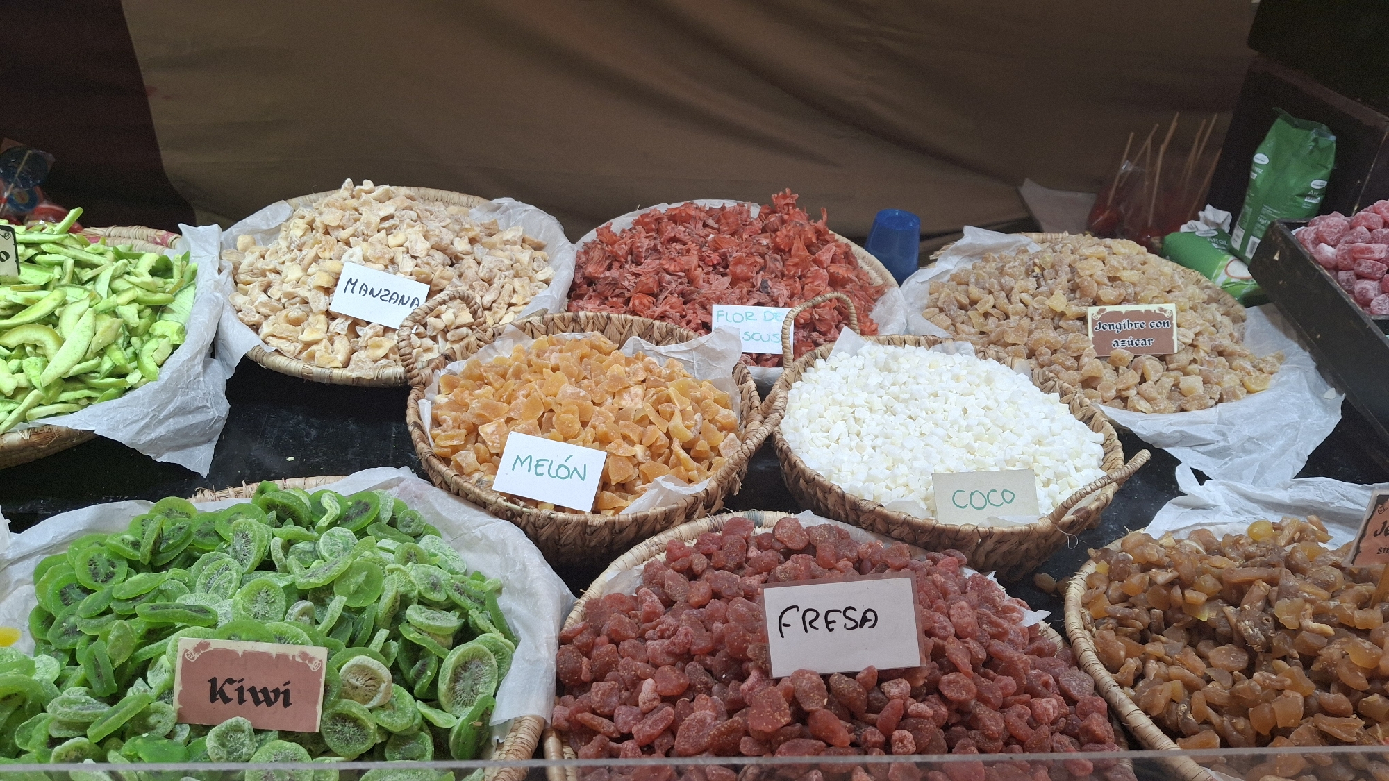 The image shows a market stall displaying a variety of dried fruits and sweets. In the foreground, a large basket is filled with green, sliced kiwi fruit. To its right, a basket holds small, red dried strawberries, labeled "FRESA". Further to the right, a basket overflows with amber-colored dried fruit, likely apricots or similar. In the middle ground, from left to right, are baskets containing: pale yellow dried apple slices labeled "MANZANA", a pile of red dried hibiscus flowers labeled "FLOR DE SCUS", orange dried melon pieces labeled "MELON", and a heap of small white diced fruit, likely coconut, labeled "COCO". Behind the melon, a basket is filled with light brown pieces of candied ginger labeled "Jengibre con azucar". The stall appears to be set up under a brown tarp or awning. The overall scene suggests a food market in La Fresneda, Spain, likely offering a selection of traditional candied and dried fruit products.