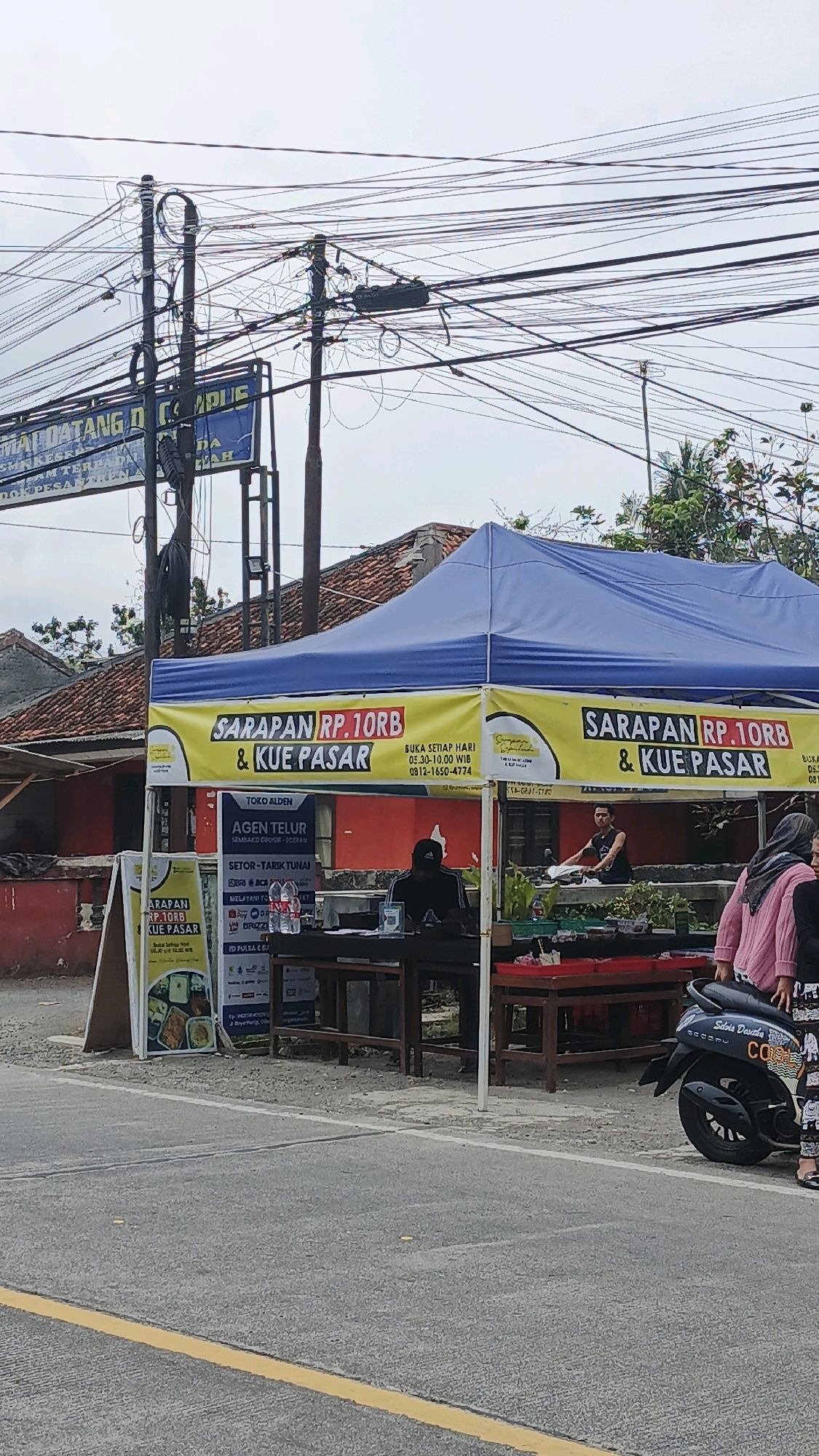 A roadside food stall with a blue tent is set up in Cibenda, Indonesia. The tent has yellow banners that advertise "SARAPAN RP. 10RB & KUE PASAR" which translates to "Breakfast Rp. 10,000 & Market Cake." One banner indicates opening hours are 05:30-10:00 WIB (Western Indonesian Time). Under the tent, a man in a black t-shirt and cap is seated behind a table where a QR code is displayed. A woman in a pink cardigan and a woman in a patterned outfit are standing near a black motorcycle parked to the right of the stall. Another smaller sign with food images is visible to the left of the tent. In the background, a building with a red roof is partially visible. Overhead, a complex network of power lines is strung between poles. The ground is paved with asphalt and a yellow line marks the edge of the road.
