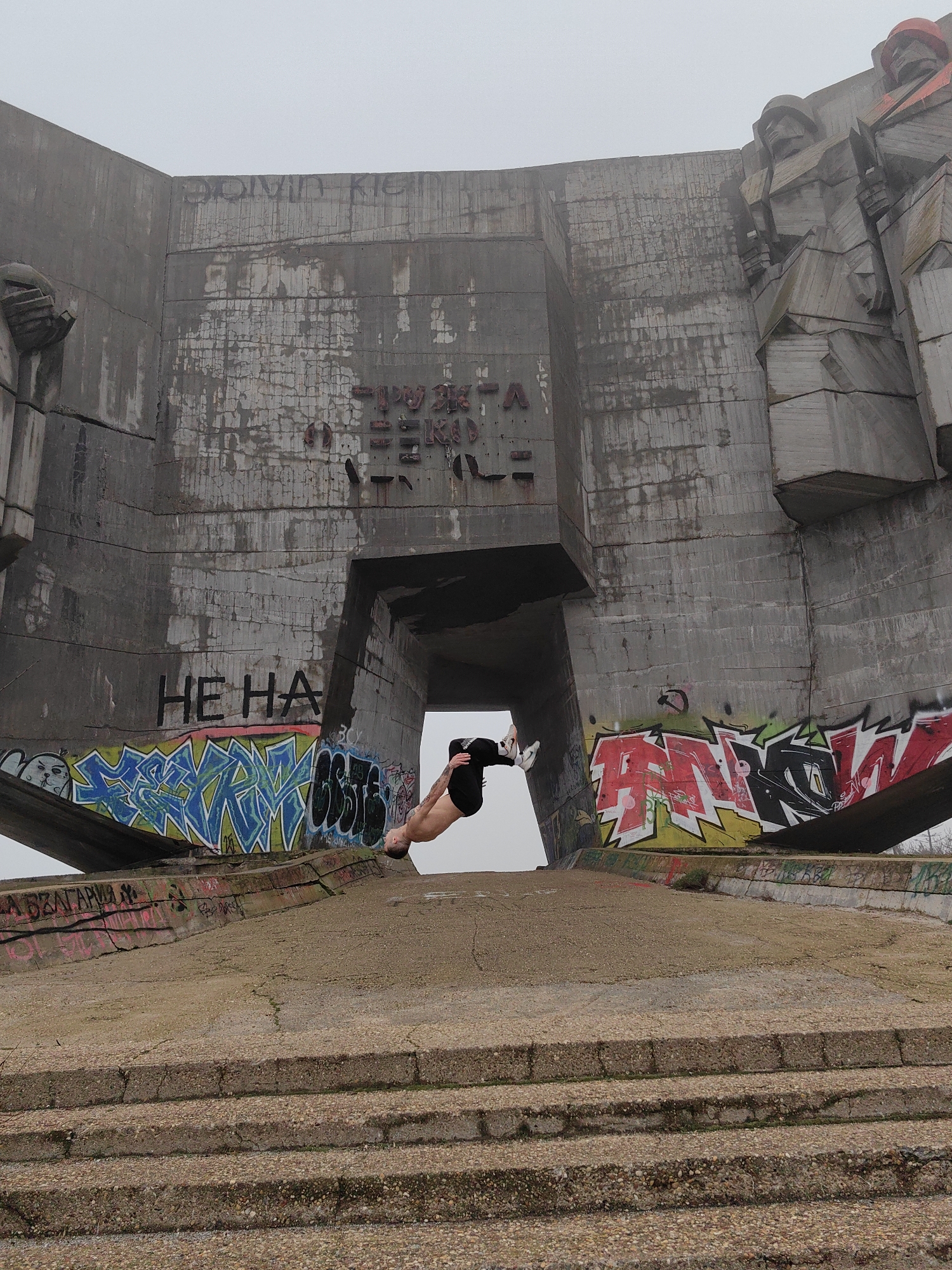 A full-length outdoor shot in Varna, Bulgaria, shows a light-skinned person doing a backflip in front of the Monument of the Bulgarian-Soviet Friendship, a large concrete structure covered in graffiti with Cyrillic text. The person is inverted, wearing black shorts and white shoes. The monument is gray and weathered, with a large opening in the center and depictions of human figures near the top right. The ground in front of the monument consists of concrete steps and a platform, also marked with graffiti. The sky is overcast.