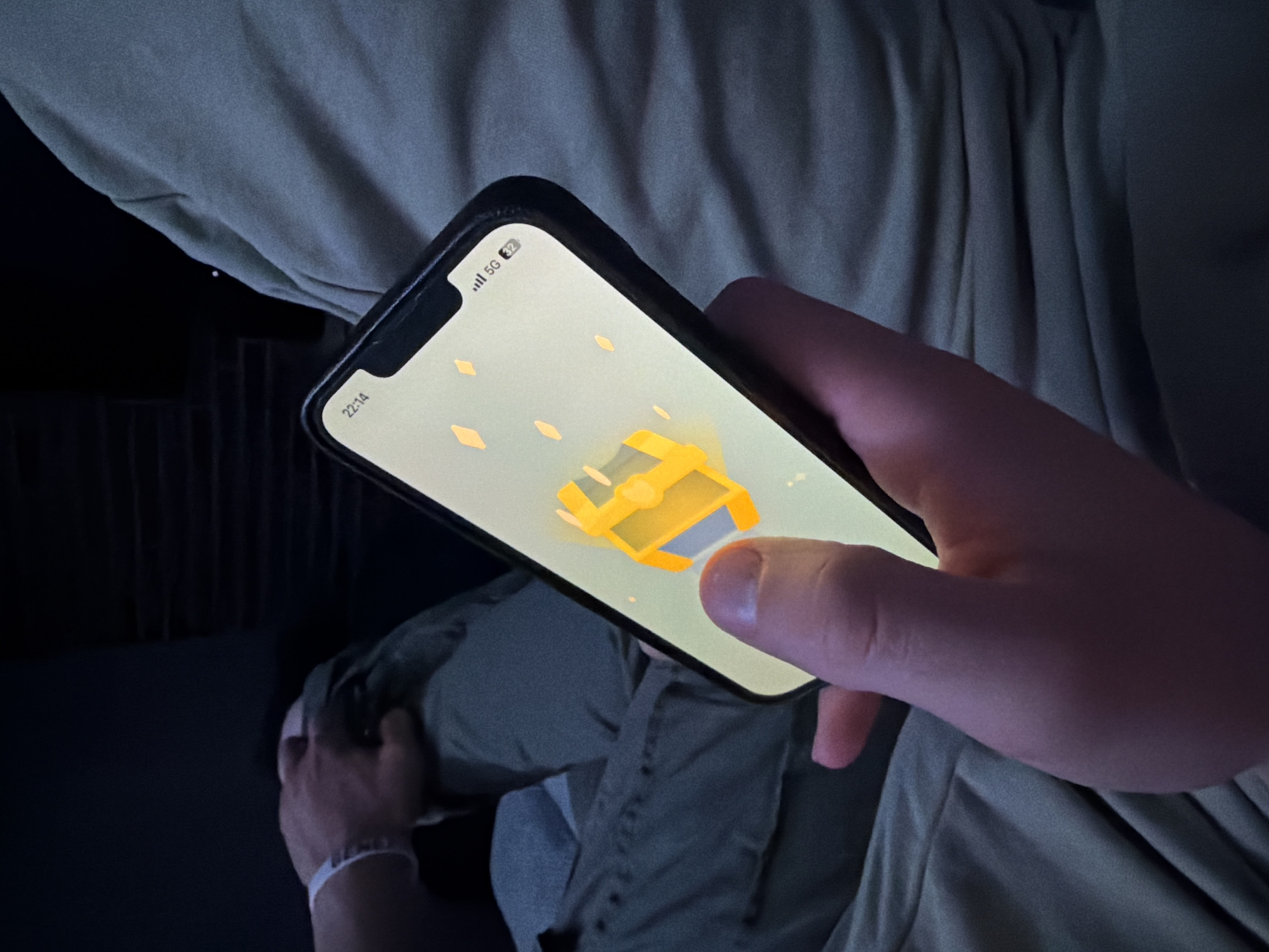 A person is holding a smartphone with a game displayed on the screen. The game appears to involve collecting yellow gems and a yellow chest. The person's finger is on the screen, presumably interacting with the game. The person is wearing what appears to be a green shirt. The background includes what looks like a bed and a wall. The phone indicates it is 22:14 and has a 5G network connection. There is no information in the image to indicate this is in the city of Buhovo, Bulgaria.