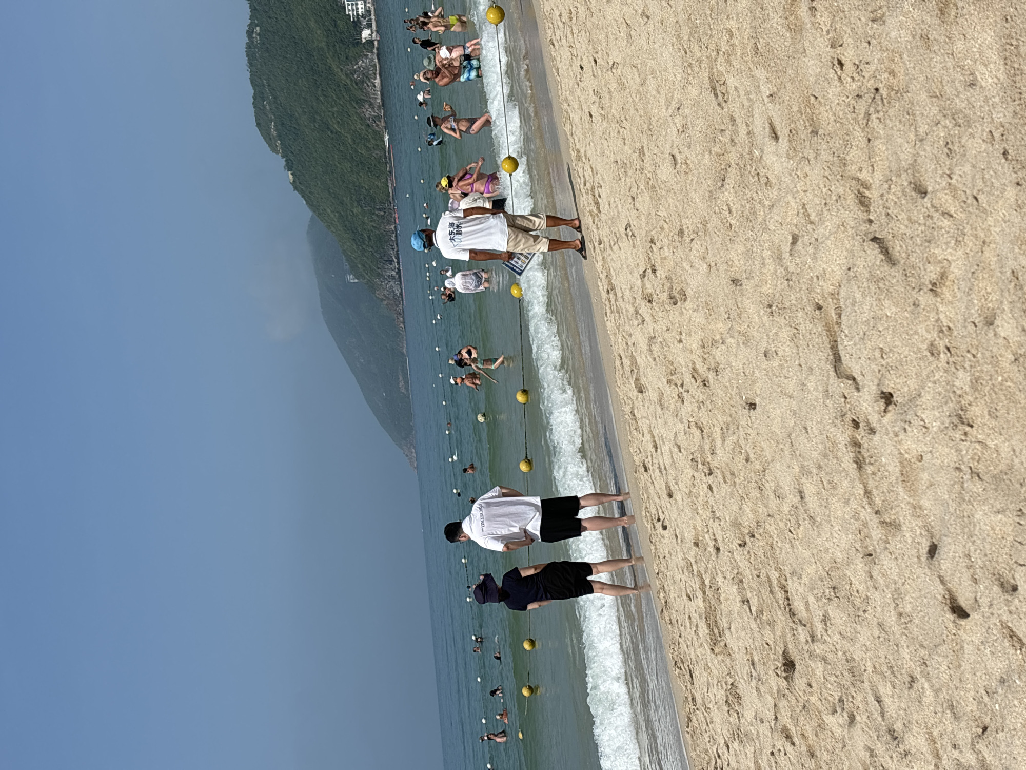 This image shows a crowded beach on a sunny day. The water is clear and blue, with gentle waves breaking on the shore. The sand is light brown and appears to be clean. In the foreground, two people are standing on the beach, facing the water. The person on the left is wearing a black hat and a black outfit, while the person on the right is wearing a white t-shirt and black shorts. Both are looking out at the ocean.
In the middle ground, there are many people swimming and playing in the water. Some are wearing swimsuits, while others are wearing casual clothing. There are also several yellow buoys in the water, which may mark a swimming area or a safety zone. In the background, there are green, tree-covered hills that slope down to the sea. The sky is clear and blue.
A man wearing a white t-shirt with Chinese characters, khaki shorts, and a blue cap is standing on the beach, facing the water, and holding a pamphlet. He appears to be a staff member, possibly a lifeguard or information attendant. The beach is located in Jiyang District, China.