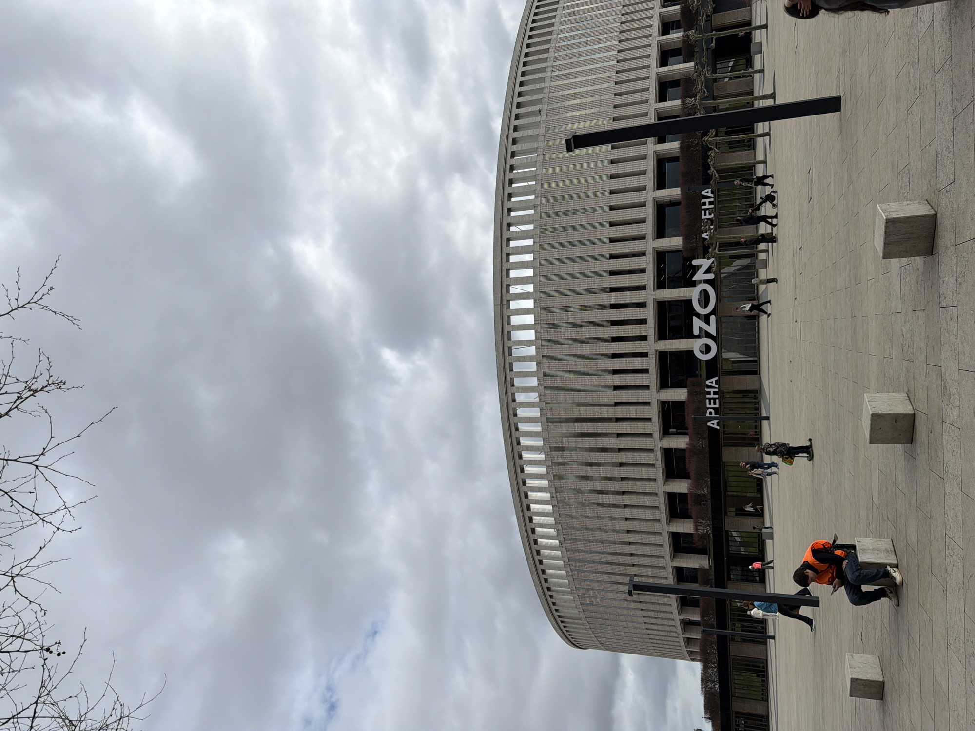 The image depicts the exterior of the "Arena OZON" building in Krasnodar, Russia, under a cloudy sky. The building has a curved facade with vertical lines and rectangular windows. The "OZON" logo is displayed in large white letters on the building's exterior, with "APEHA" written above it. In front of the building, there is a paved plaza with several concrete cubes placed around. Several people are walking or standing on the plaza. One person is sitting on a concrete cube, wearing an orange vest and using a device that looks like a phone. Some bare trees are visible in the left foreground.