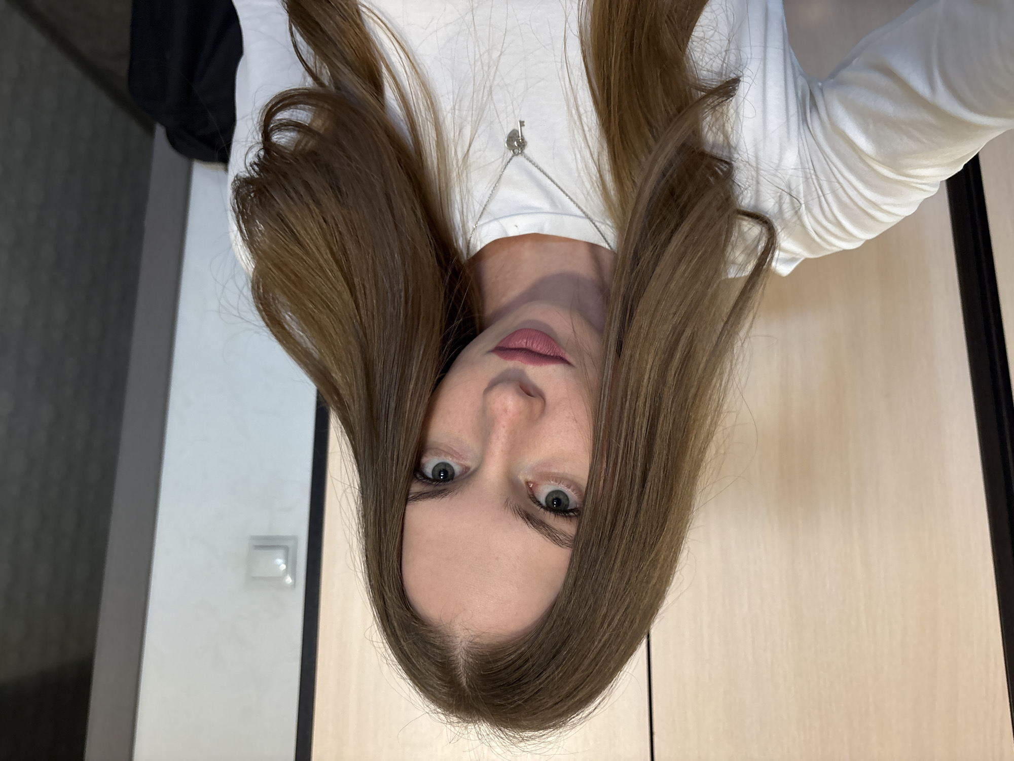 A young woman with long brown hair and light skin is taking a selfie. She is wearing a white long-sleeved shirt and a silver necklace with a heart-shaped pendant and a key. She has light green eyes and her lips are painted with a matte pink lipstick. The background appears to be an interior, possibly a closet or room with light-colored wooden doors and a light switch on the wall. The image does not contain any information that can be used to determine its location in the city of Perm, Russia.