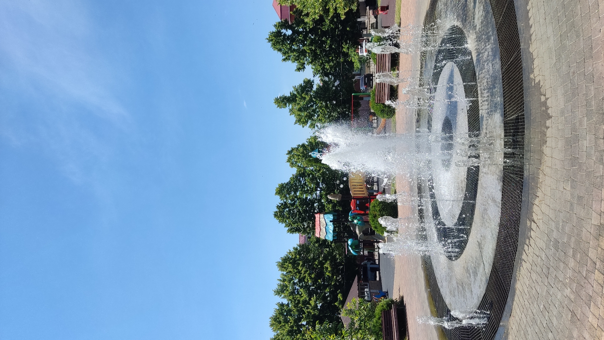 A wide shot captures a circular fountain in the foreground, with water jets spraying upwards and outwards in a lively display. The fountain is set within a paved area of interlocking bricks, surrounded by a metal grate. In the background, a row of lush green trees forms a backdrop, with glimpses of buildings and a playground peeking through the foliage. The sky above is a clear, bright blue with a few wispy clouds. Several park benches are visible, some occupied by people, suggesting a public space. A couple of cars are parked nearby. The scene appears to be a well-maintained public park in Takhtamukai, Russia, possibly on a sunny day.