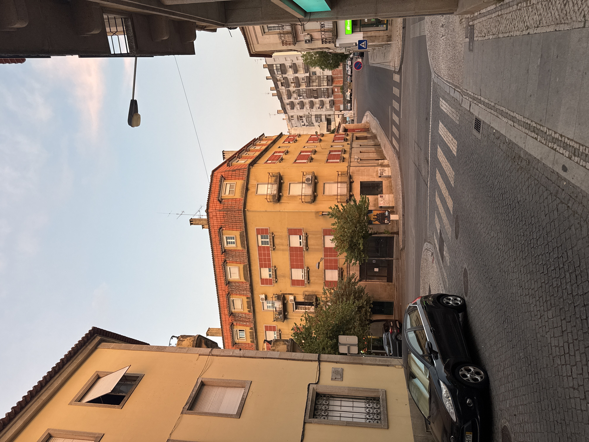 The image depicts a street corner in Castelo Branco, Portugal, at dusk. A bright yellow building with a red-tiled roof dominates the left side of the frame. It features multiple windows with red shutters and balconies. A dark-colored hatchback car is parked on the cobblestone street in the foreground. Across the street, another yellow building with a red-tiled roof is visible, along with a taller white building in the background. The sky is a soft gradient of blue and pink, indicating the end of the day. The street is paved with cobblestones and includes a pedestrian crossing. A streetlamp extends from a building on the right. The scene appears calm and quiet, with no people visible.