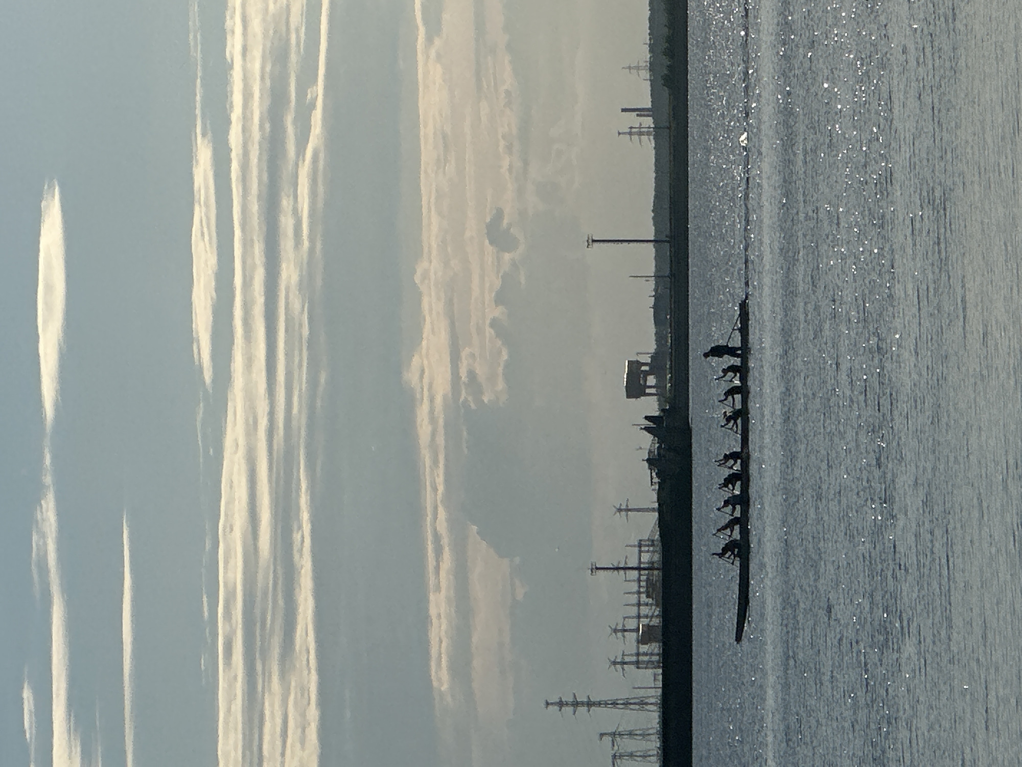A long rowing boat with at least eight occupants is silhouetted against the shimmering water of the Kama River. The rowers are engaged in the action of rowing, with oars extended and bodies leaning forward. The boat is positioned in the middle of the river, with a distant shoreline visible. On the shoreline, a dark industrial silhouette is apparent, featuring tall transmission towers and various structures, suggesting an industrial or power generation facility. The sky above is a pale blue, marked by horizontal bands of light and dark clouds, with the sun's rays creating highlights on the water's surface. The scene is a daytime view, likely during a calm period with minimal waves on the river. The location is the Kama River in Chaikovsky, Russia, a city known for its industrial presence and its location on the Kama River.
