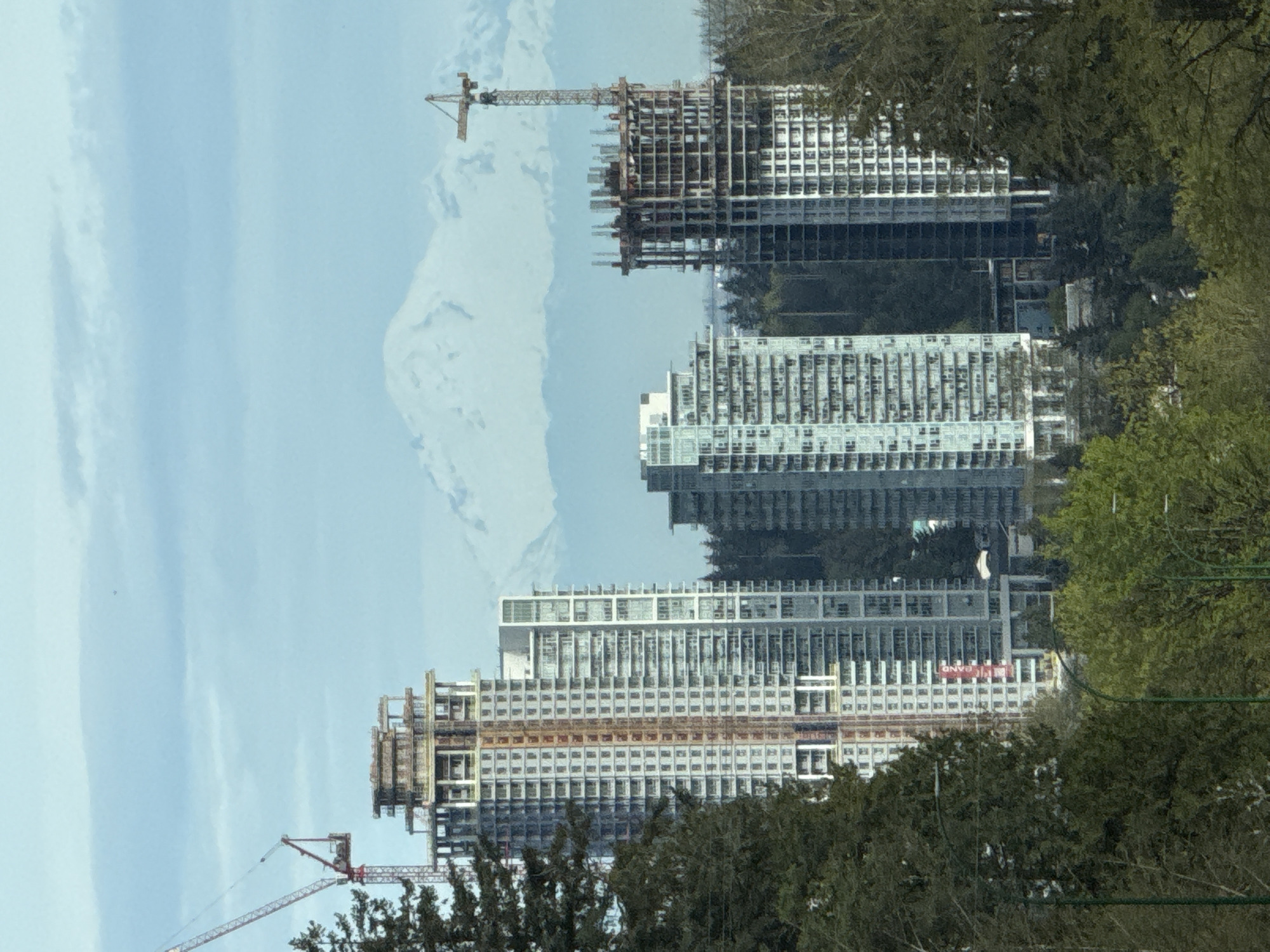 The image depicts a view of several high-rise buildings under construction in Burnaby, Canada. In the foreground, trees partially obscure the view. Three completed high-rise buildings are visible. Construction cranes are adjacent to partially constructed buildings. The sky is partly cloudy.