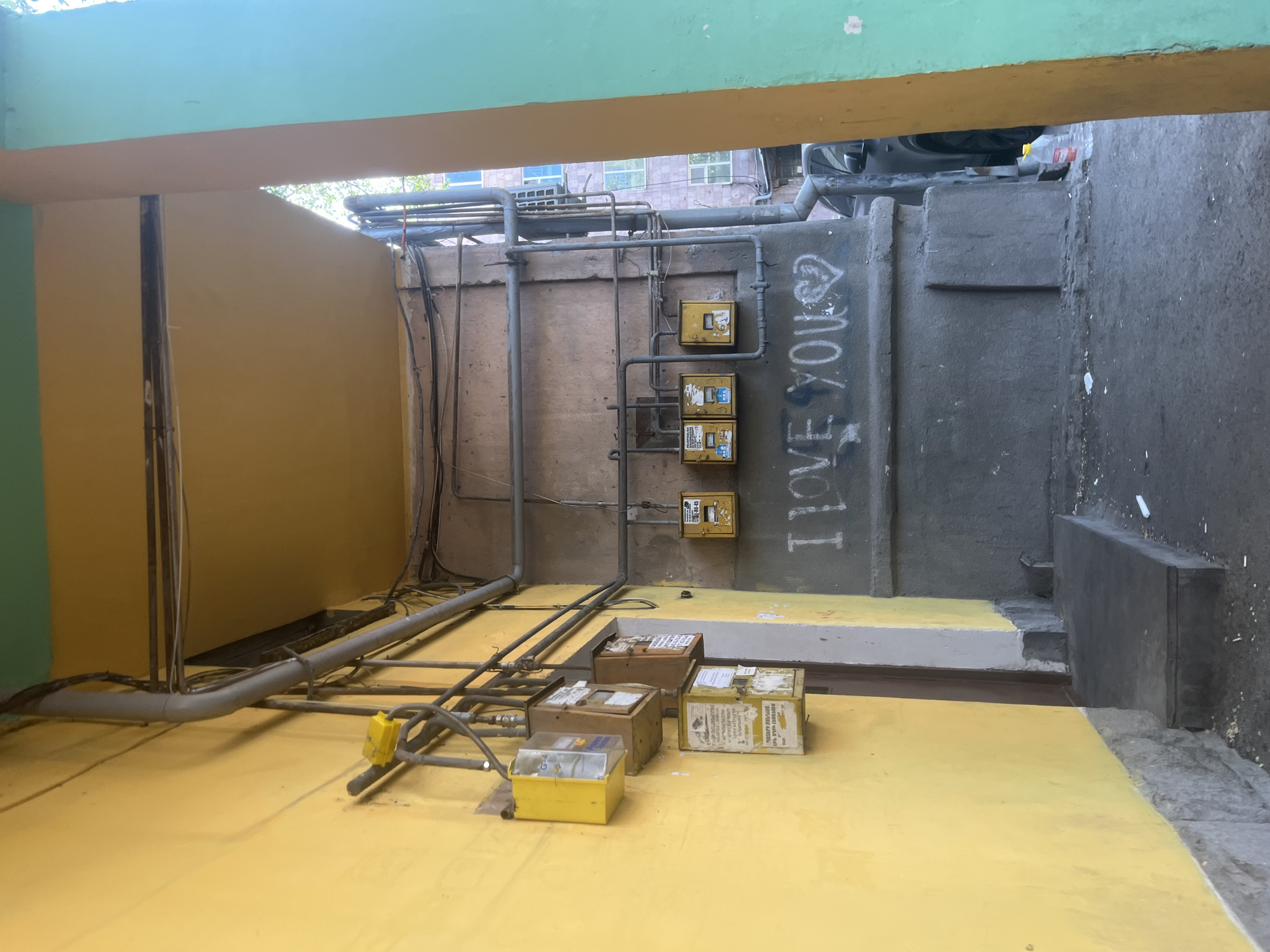 The image depicts a utility area, possibly an alleyway or storage space, in Yerevan, Armenia. The walls are painted in a combination of green, yellow, and grey. There are various utility installations visible, including gas or water meters mounted on the wall and an array of pipes and electrical cables running along the wall and floor. Several yellow-painted boxes with visible labels and markings are placed on the floor. Graffiti, including the words "I LOVE YOU," is sprayed on one of the walls. The overall setting appears somewhat neglected, with signs of weathering and general wear.