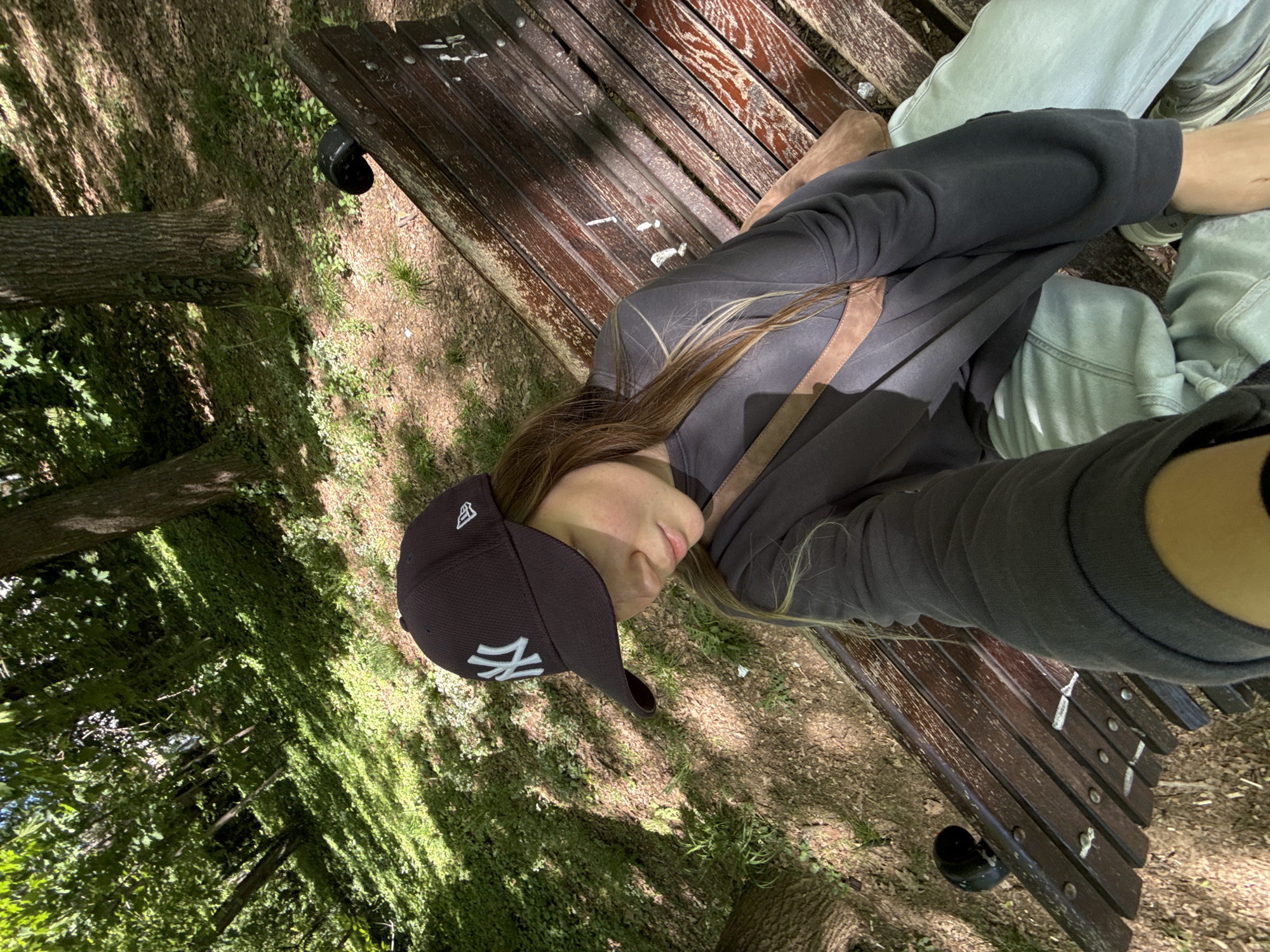 A young woman wearing a gray sweatshirt and a black baseball cap with a New York Yankees logo is lying on a wooden park bench. Her head is tilted to the left, and her face is partially obscured by the brim of the cap. The bench is positioned in a park with trees, grass, and dappled sunlight. The location is Vozdovac Urban Municipality, Serbia.